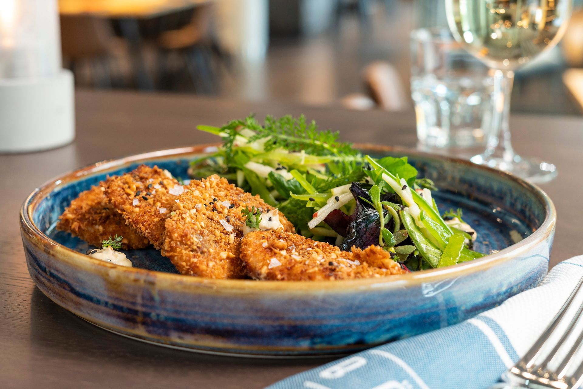 Ein Teller mit goldbraun panierten Schnitzeln, serviert mit einem frischen grünen Salat auf einem dunklen Tisch neben einem Glas Weißwein.