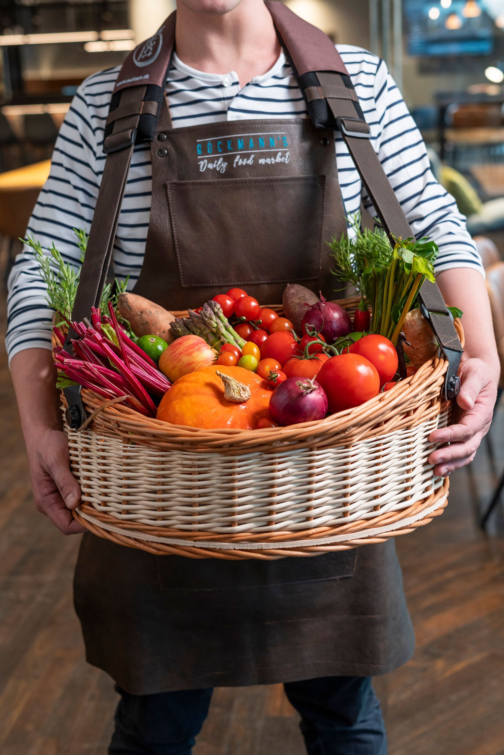 Eine Person mit brauner Schürze und gestreiftem Hemd hält einen großen Weidenkorb, der mit buntem, frischem Gemüse gefüllt ist.