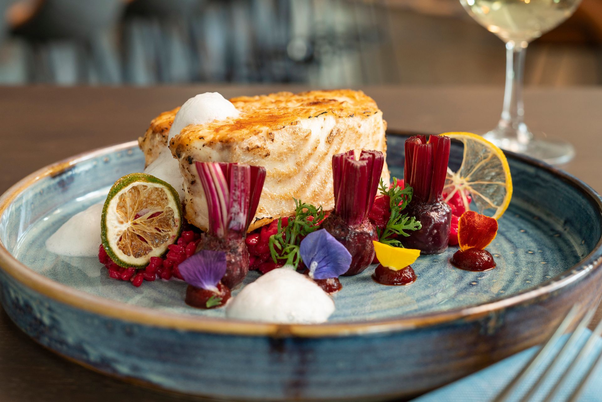 Ein angebratenes Weißfischfilet, serviert auf einem blauen Keramikteller mit gerösteten Rote-Bete-Scheiben, essbaren Blüten und Zitronenscheiben.