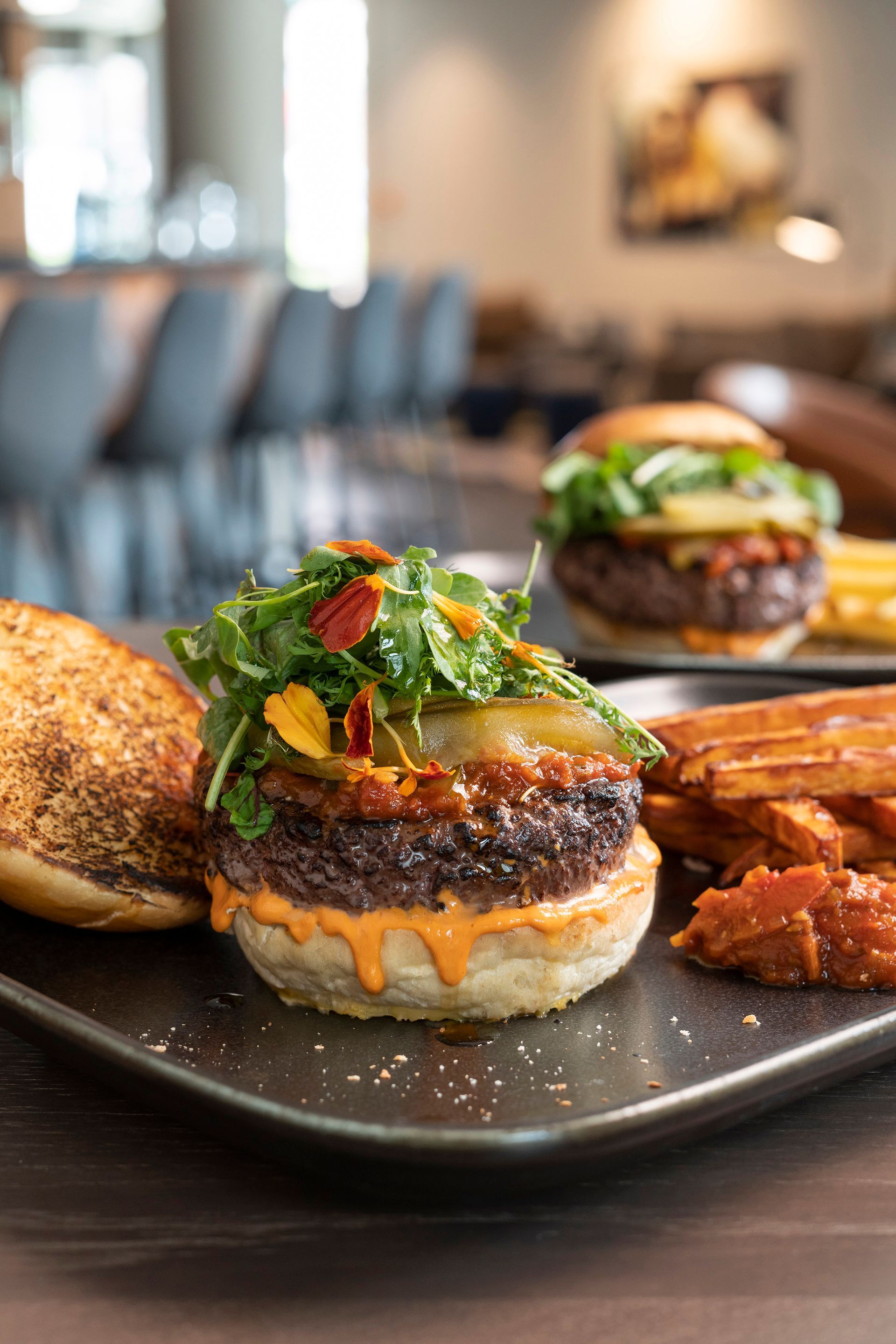 Zwei Gourmet-Burger mit Salat und roter Soße, serviert mit Kartoffelecken auf einem dunklen Teller im Restaurantambiente.