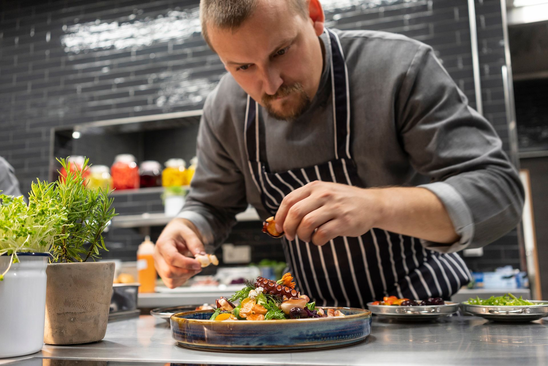 Ein Koch in grauem Hemd und gestreifter Schürze garniert sorgfältig ein Gericht auf der Arbeitsfläche einer modernen, dunkel gefliesten Küche.