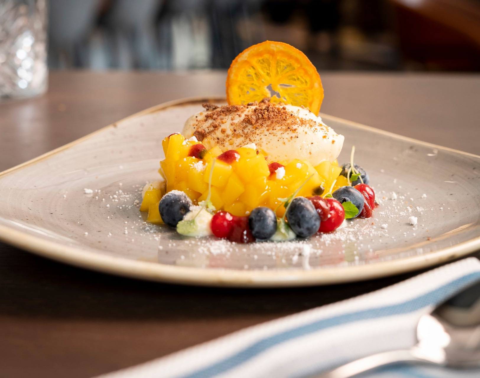 Ein auf einem Teller angerichtetes Dessert mit einer Kugel Sorbet, garniert mit Zitrusfrüchten, umgeben von gewürfelten Mangos und frischen Beeren.