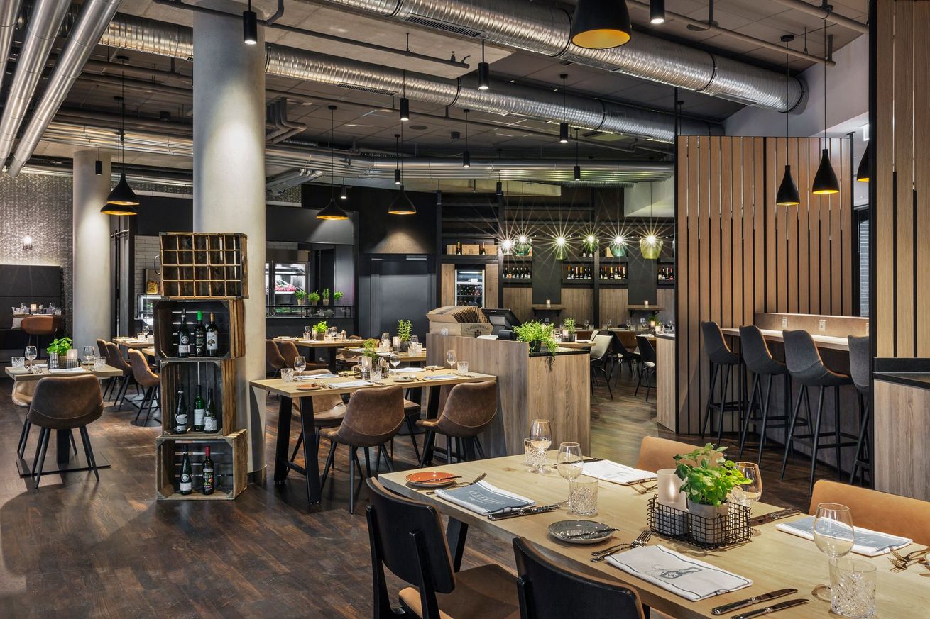 Modernes Restaurantinterieur mit Holztischen, Stühlen, einem Weinregal und Pendelleuchten in dunklem, elegantem Dekor.
