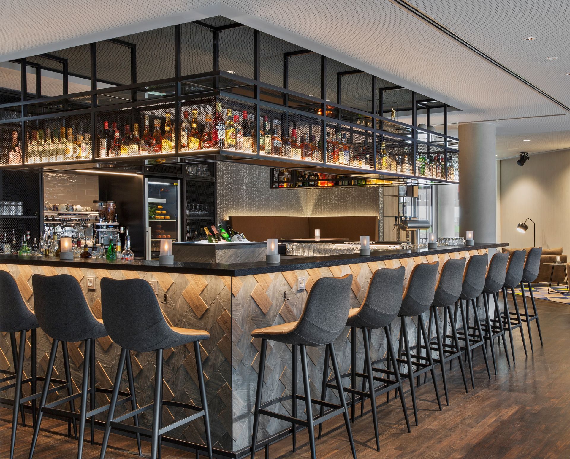 Eine moderne Bar mit dunklem Holzboden, Theke im Fischgrätmuster und einem hinterleuchteten Metallregal voller Spirituosenflaschen.