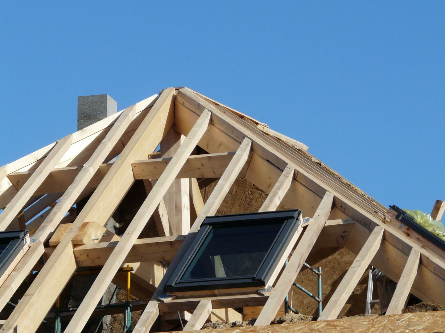 Charpente de toit en bois en construction, avec une fenêtre de toit installée sur fond de ciel bleu dégagé.