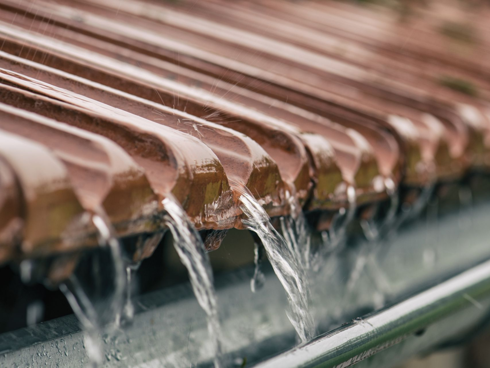 De l'eau de pluie s'écoule d'un toit de tuiles brunes dans une gouttière métallique.