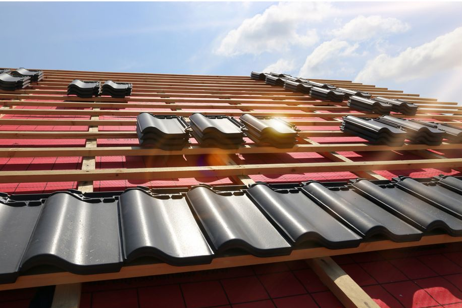 Des tuiles noires sont posées sur un toit en pente aux lattes de bois apparentes, sous un ciel lumineux.