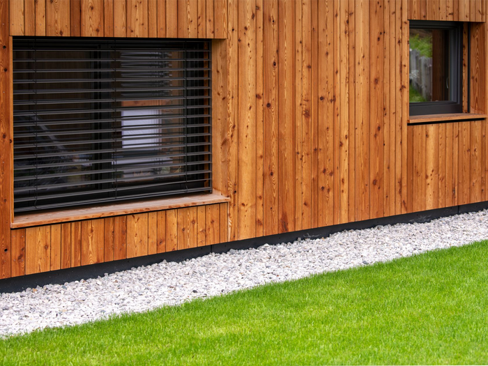 Une maison moderne avec un bardage vertical en bois., une grande fenêtre avec des stores noirs et une bordure de gravier.