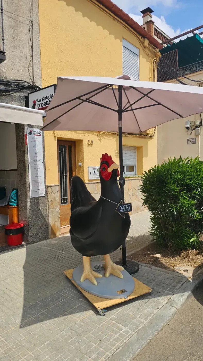 Estatua de pollo negro bajo un paraguas blanco, en una acera frente a un edificio amarillo.
