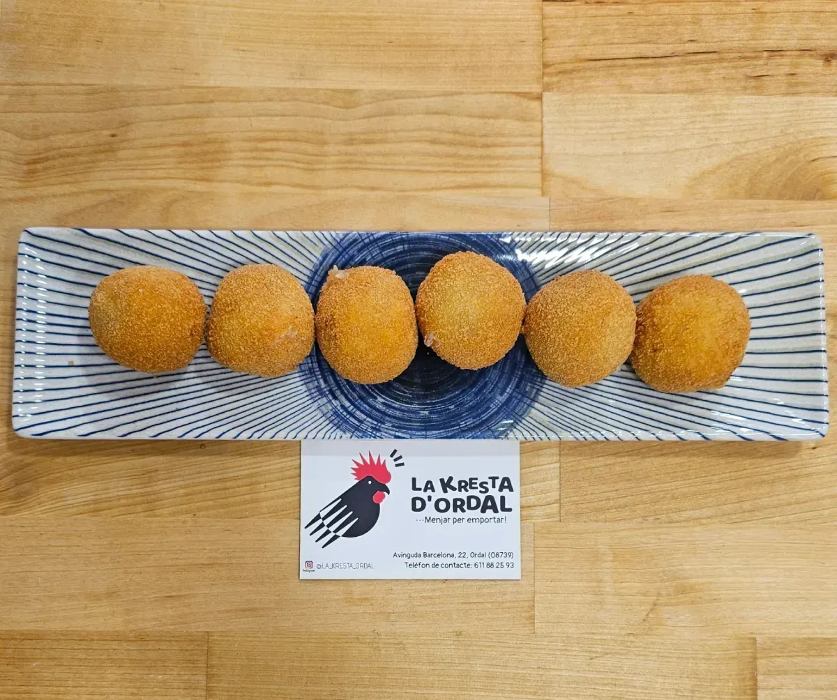 Seis croquetas doradas en un plato rectangular estampado, sobre una superficie de madera. Se ve un logotipo debajo.