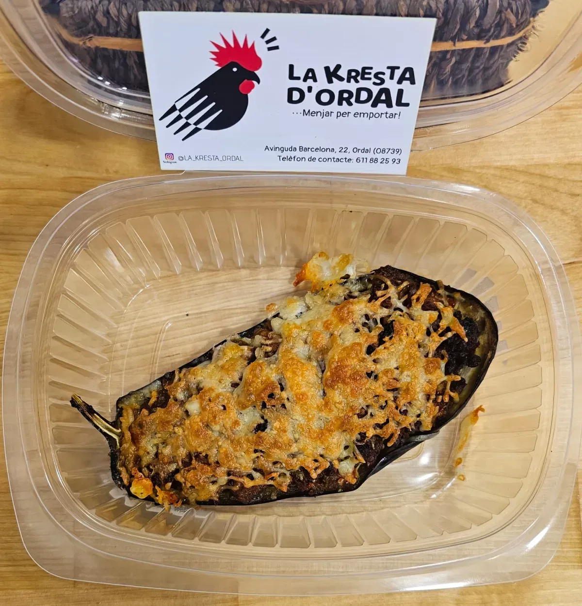 Plato de berenjenas al horno en recipiente de plástico, con una tarjeta con el logotipo de un gallo en el fondo.