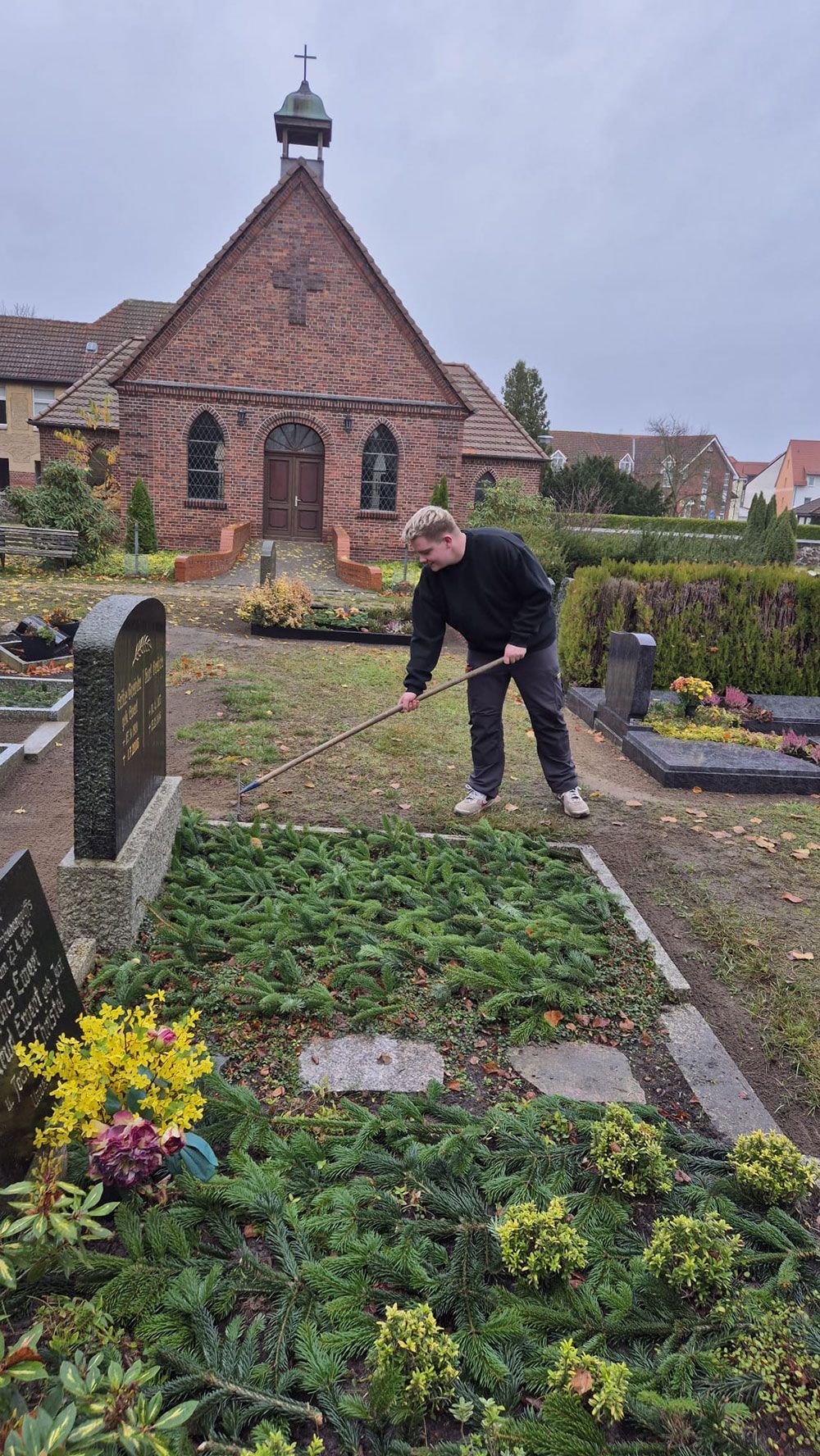 Dekoratives Eisenkreuz im Vordergrund, im Hintergrund ein verschwommenes, farbenfrohes Blumendenkmal.