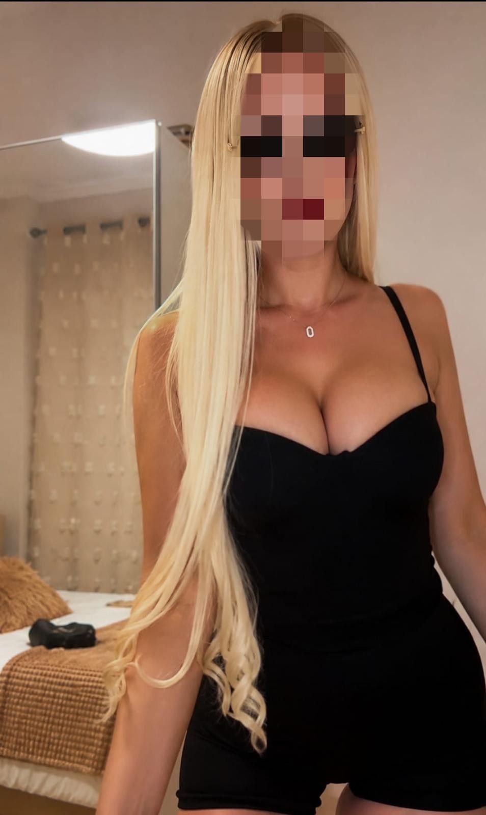 Mujer rubia con vestido negro de pie en el interior de una casa, cerca de una silla de mimbre y cortinas beige.