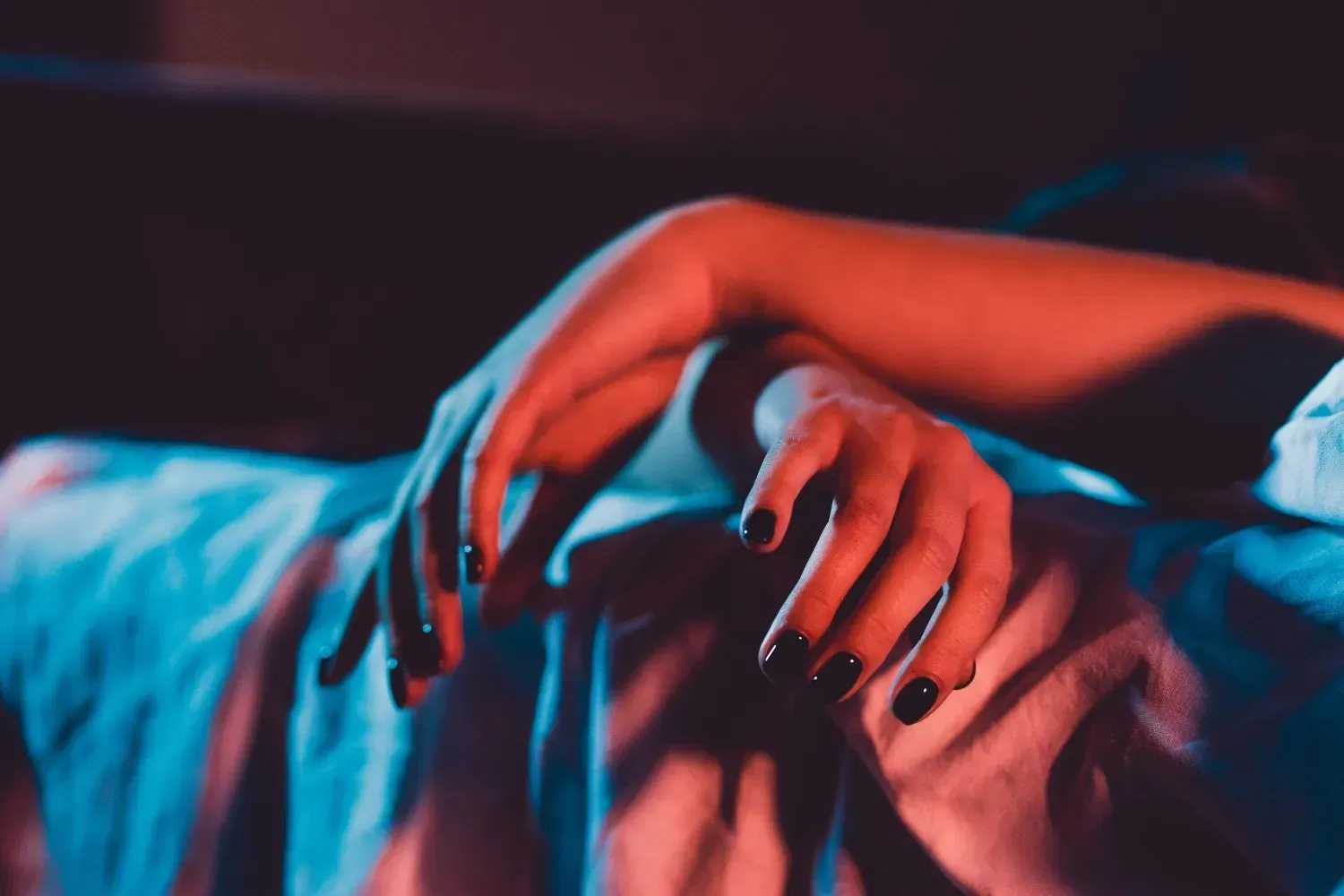Manos con esmalte de uñas oscuro descansando sobre una cama iluminada con luz azul en una escena tenue y melancólica.