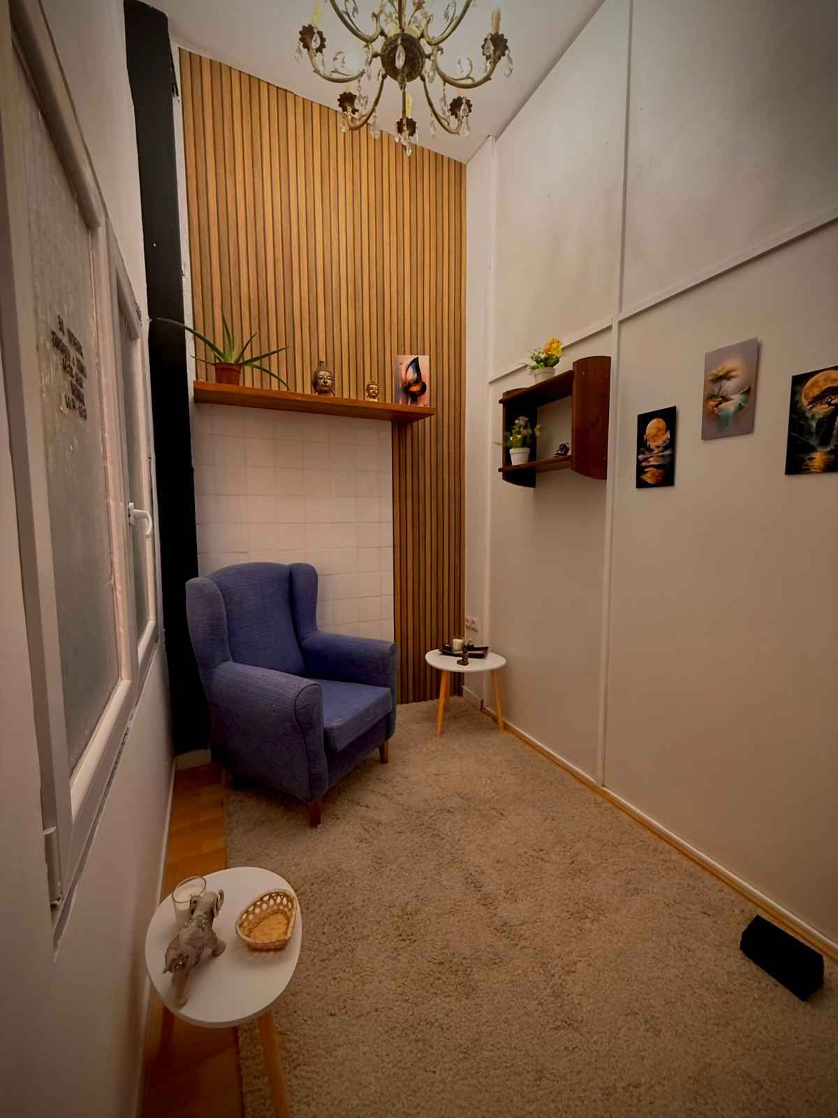 Pasillo estrecho con un sillón azul, cuadros en la pared, una lámpara de araña y una ventana alta con cortinas.
