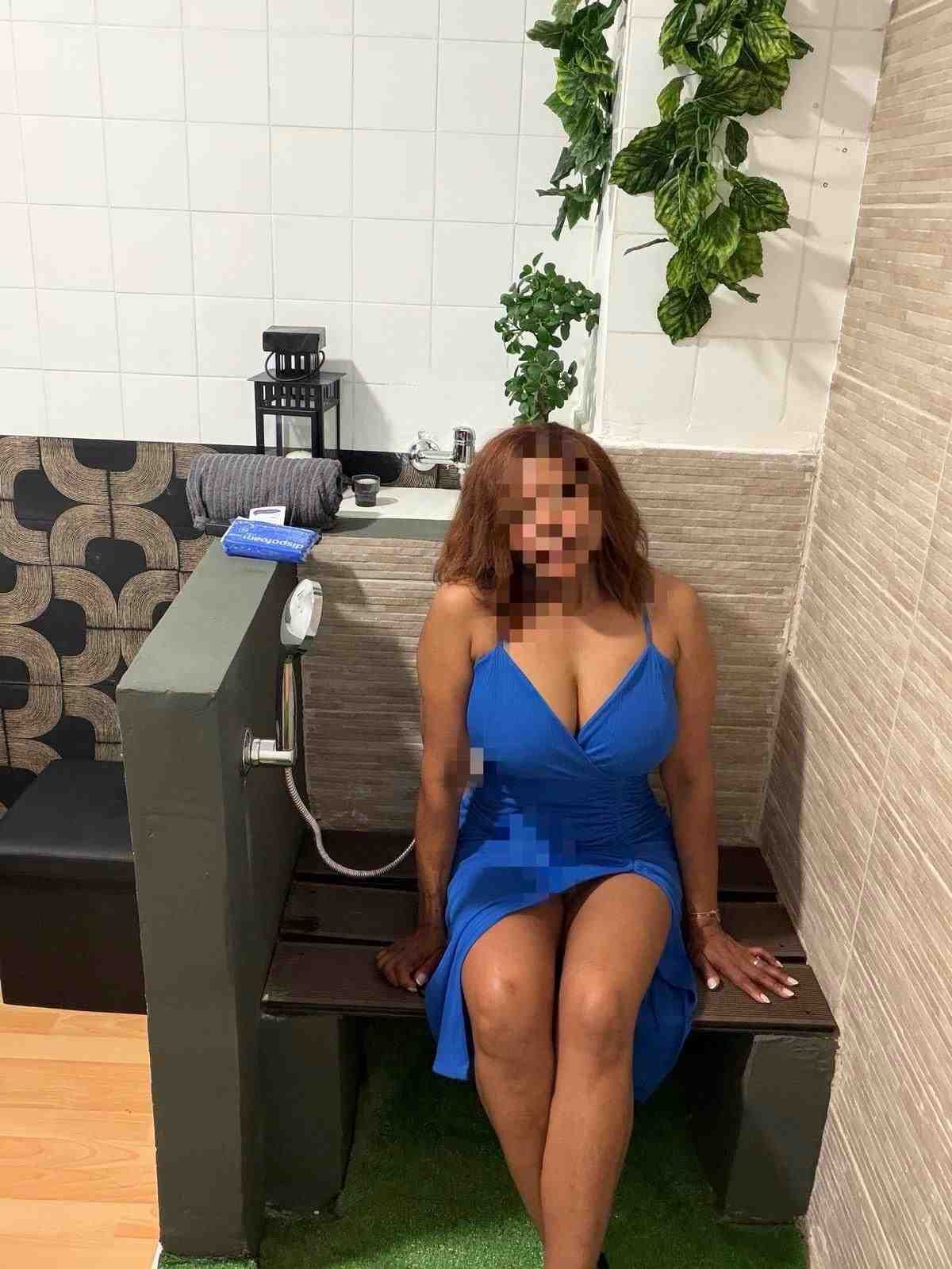 Una mujer con un vestido azul sentada en un banco en una sala de estar con suelo de baldosas y plantas.
