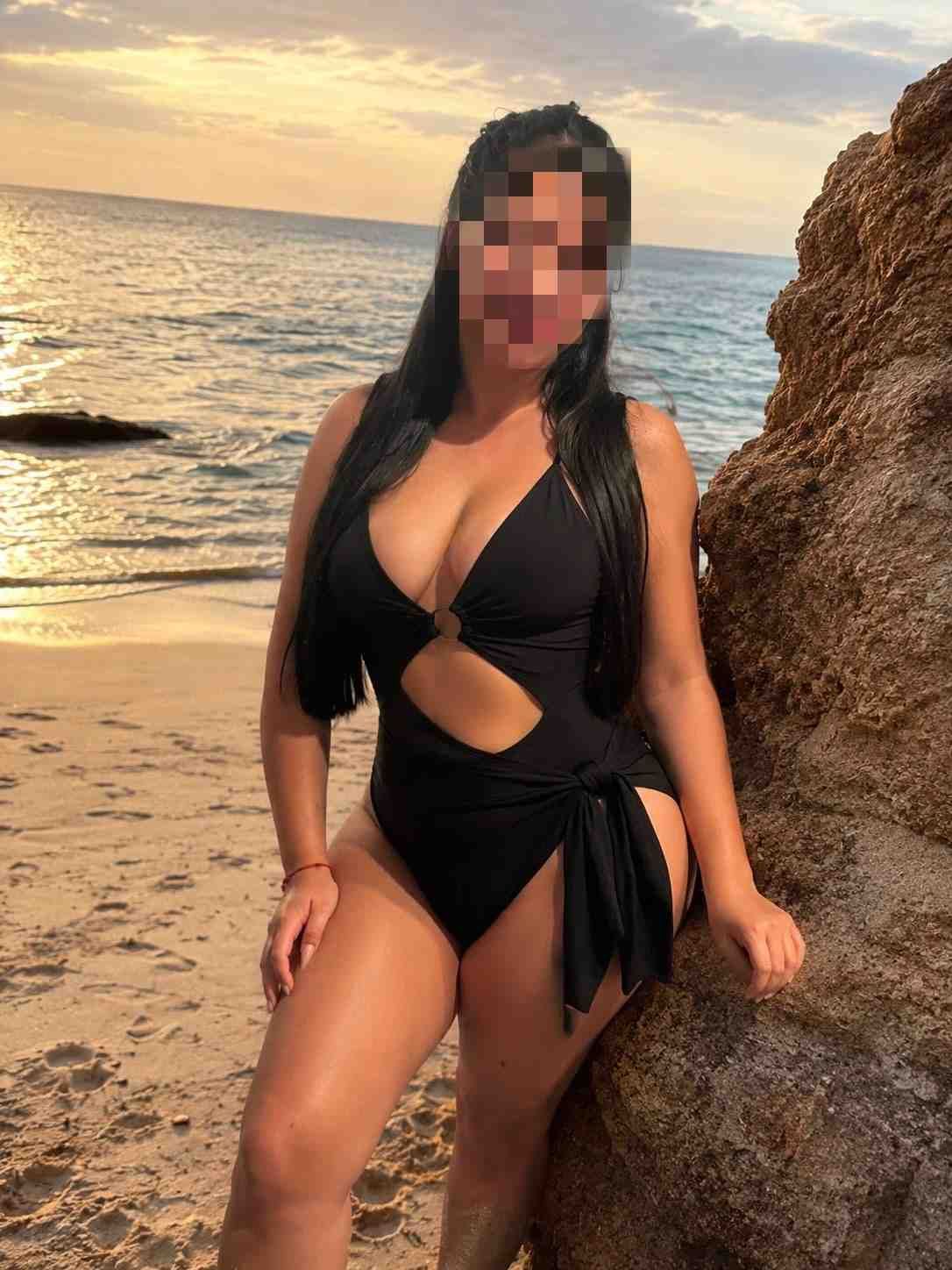 Una persona vestida de negro está de pie en una playa al atardecer, junto a una formación rocosa.