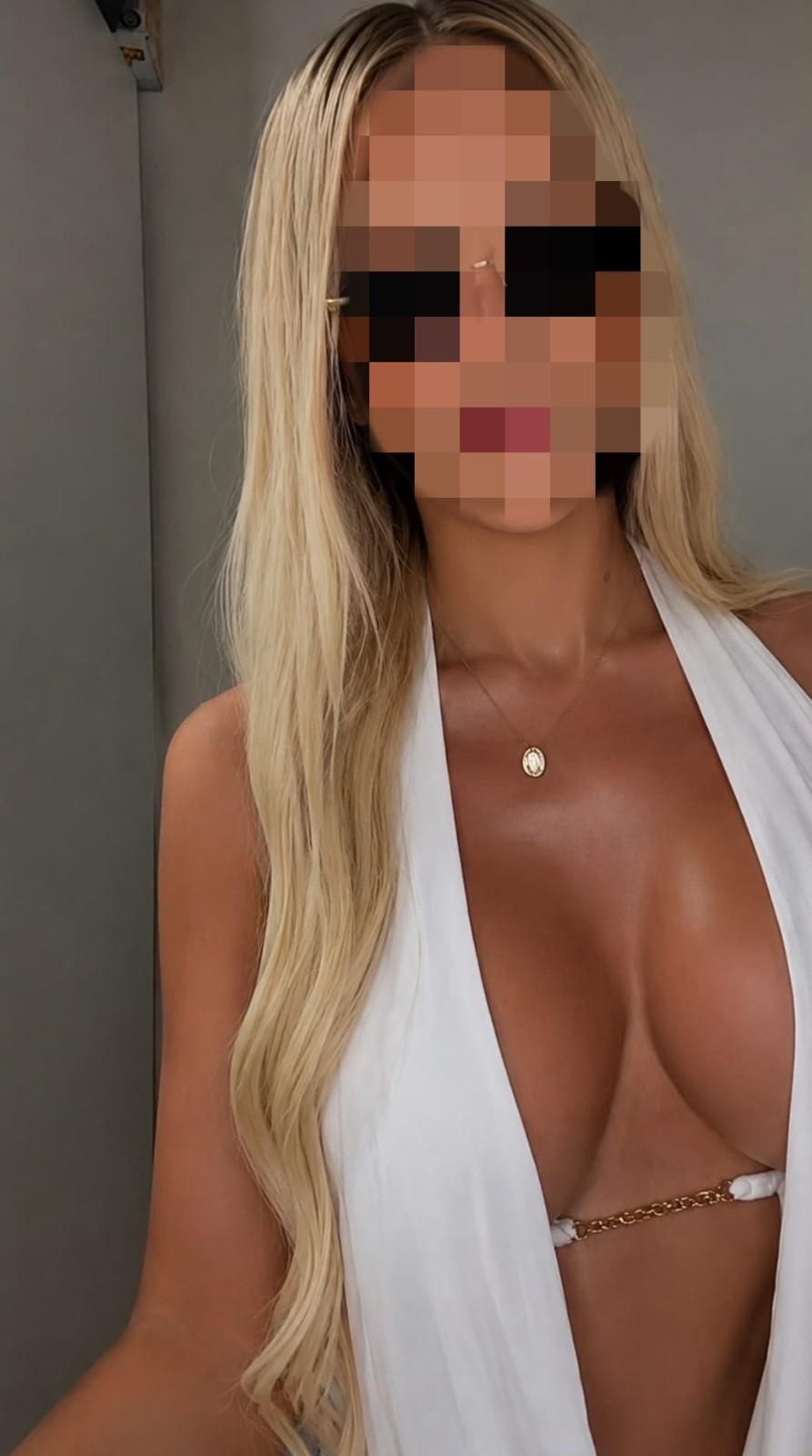 Mujer rubia con un top blanco sin tirantes posando sobre un fondo oscuro.
