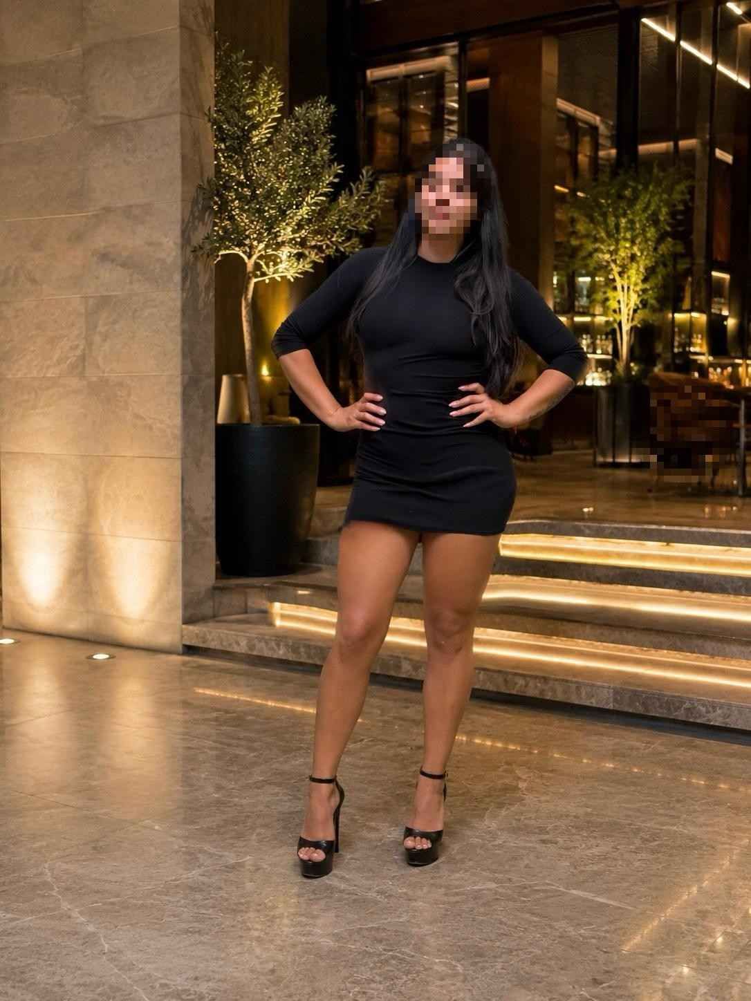 Mujer con vestido negro y tacones de pie frente a la entrada iluminada de un edificio por la noche.