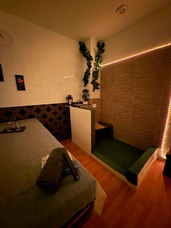 Habitación pequeña y acogedora con una cama baja, pared de azulejos, biombo de bambú, plantas y guirnaldas de luces cálidas.