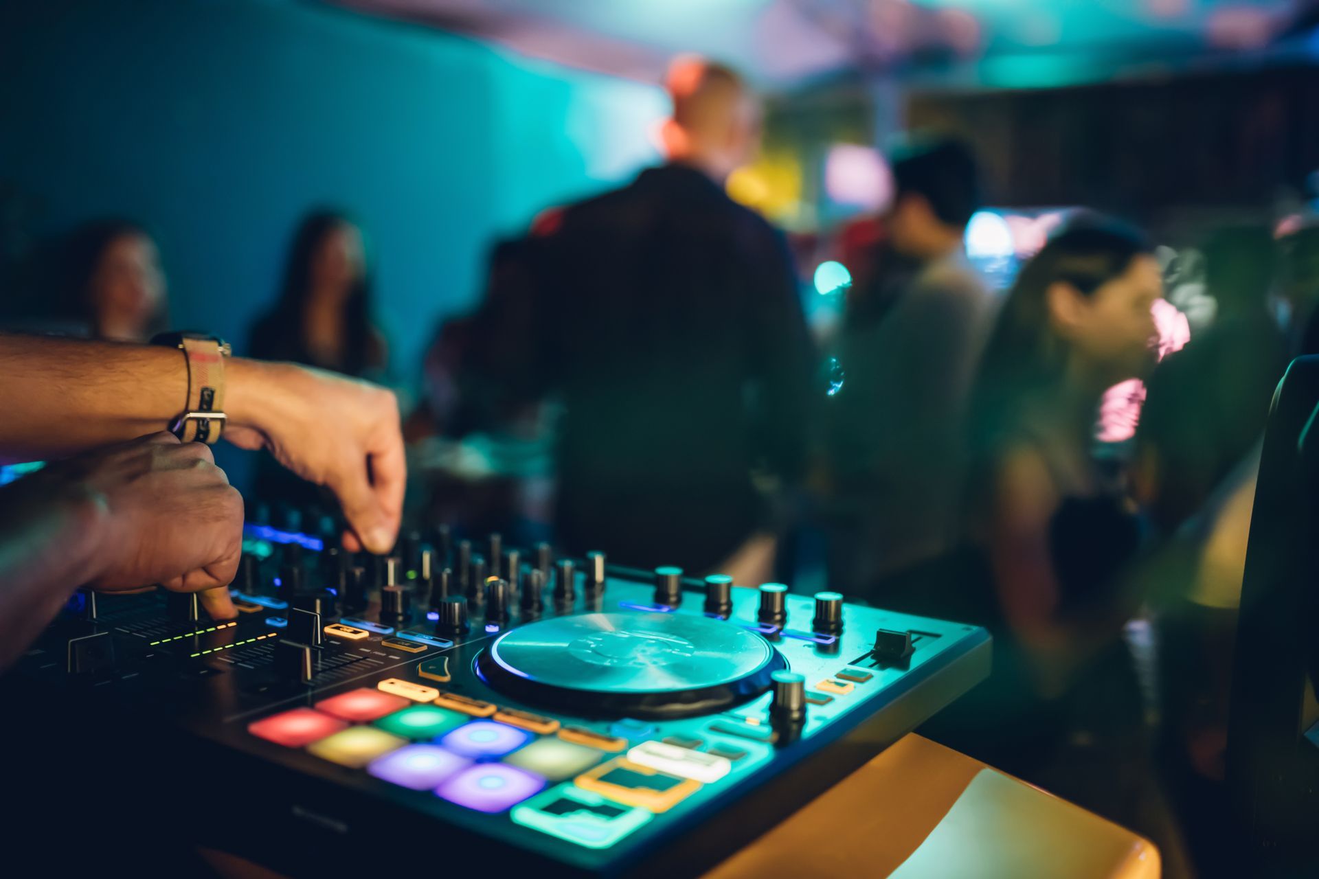Un DJ mixe de la musique sur une platine lumineuse dans une boîte de nuit, avec des lumières colorées et des danseurs en arrière-plan.