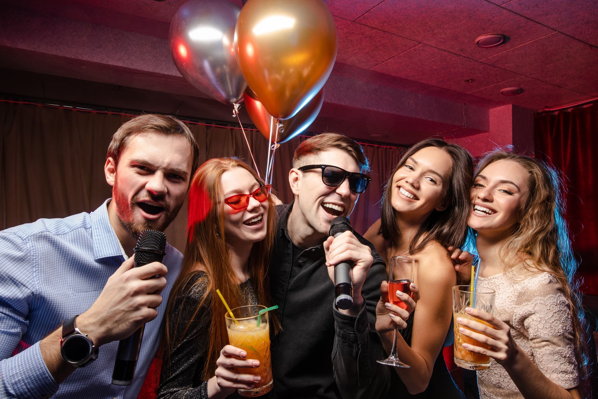 Des amis chantent et rient dans une boîte de nuit, tenant des verres et des ballons sous des lumières de fête colorées.
