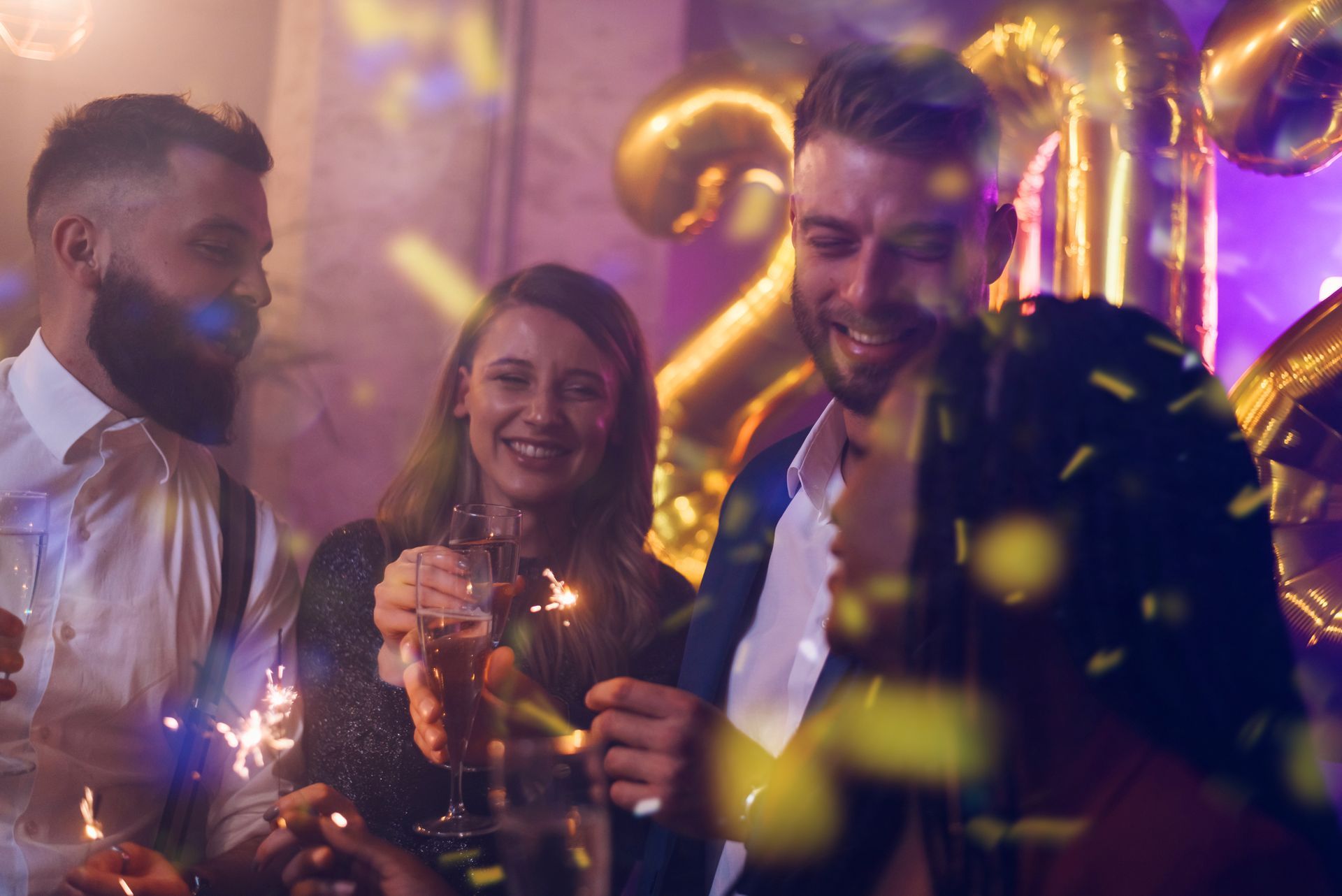 Trois amis font la fête avec des cierges magiques et des ballons dorés sous des lumières violettes.