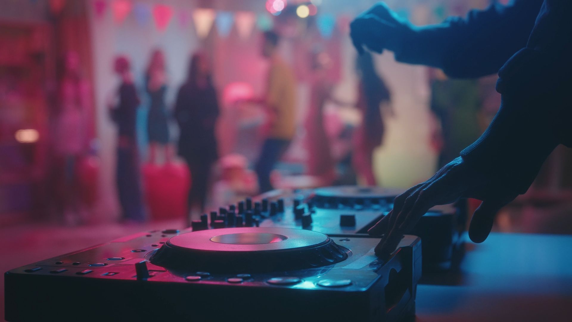Mains de DJ sur platines lors d'une soirée éclairée au néon, avec des danseurs flous en arrière-plan.
