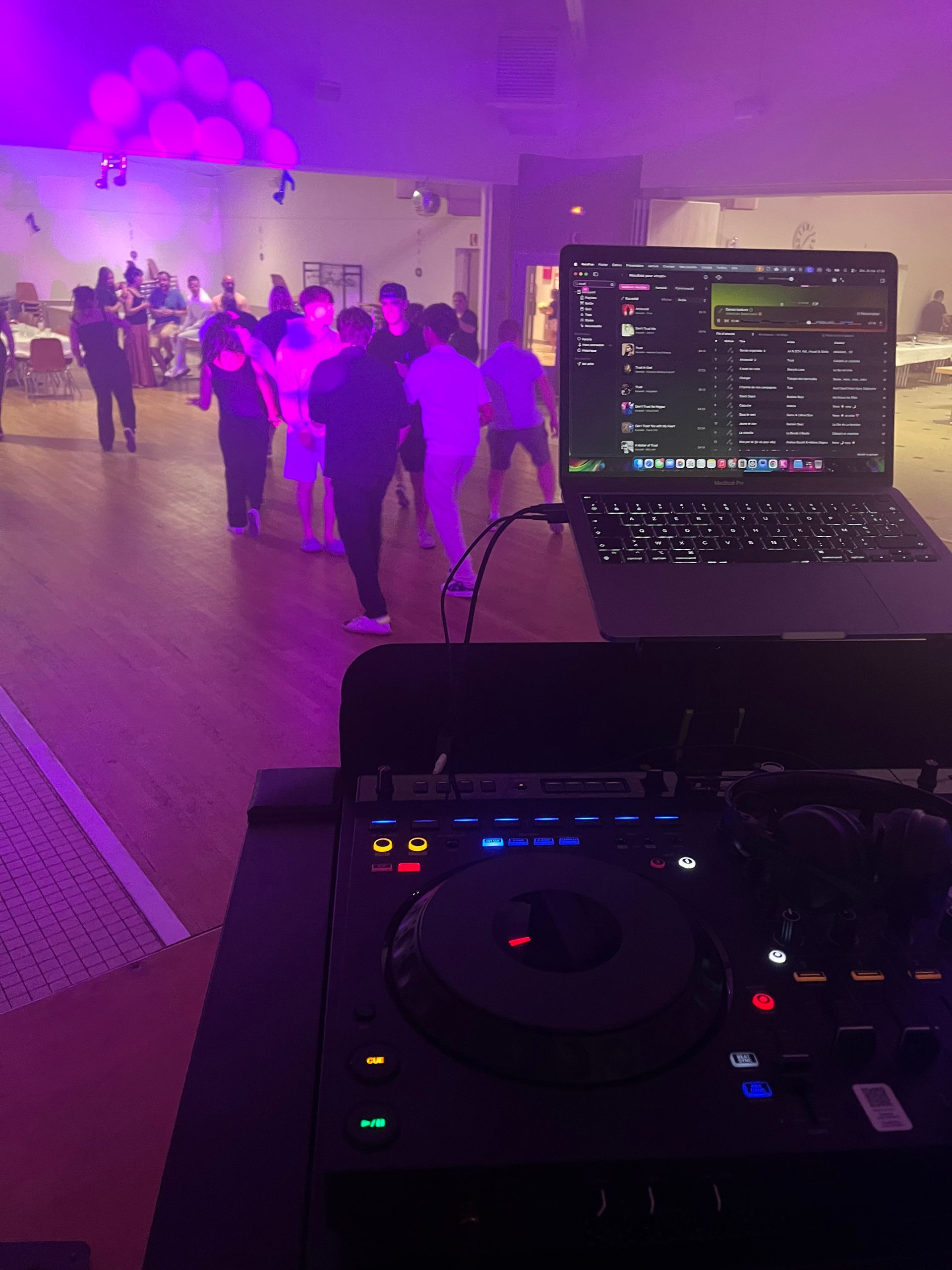 Cabine de DJ lors d'une soirée dansante éclairée en violet, avec un ordinateur portable et une table de mixage au premier plan et des personnes qui se mêlent en arrière-plan.