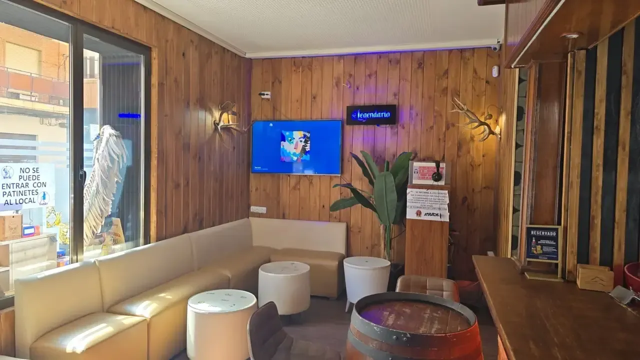 Interior de un bar con paneles de madera, un televisor, un sofá, taburetes redondos blancos y una mesa de barril de madera.