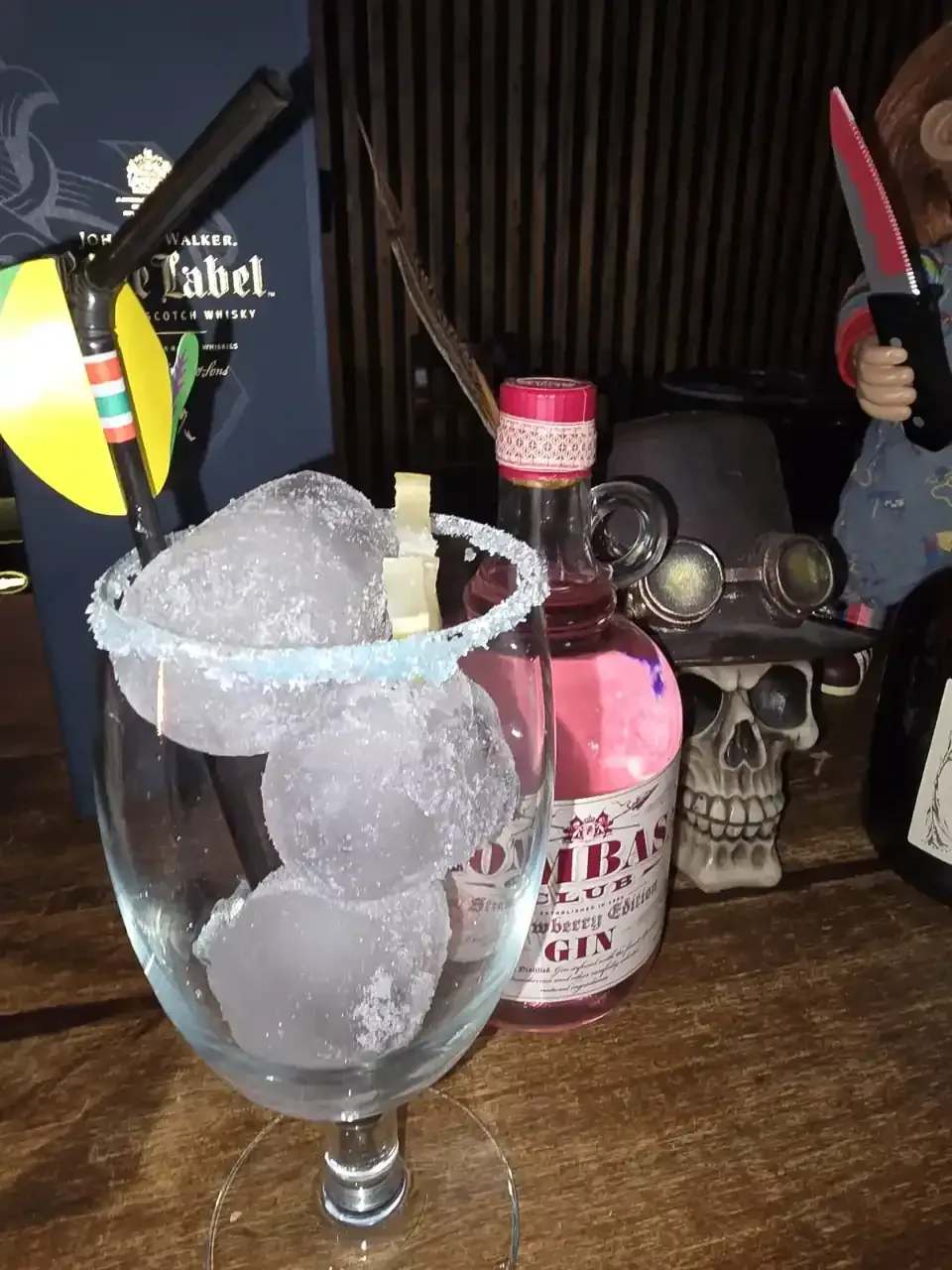 Copa de cóctel con hielo, borde de azúcar y decoraciones; botella de ginebra rosa, calavera y muñeco Chucky en el fondo.