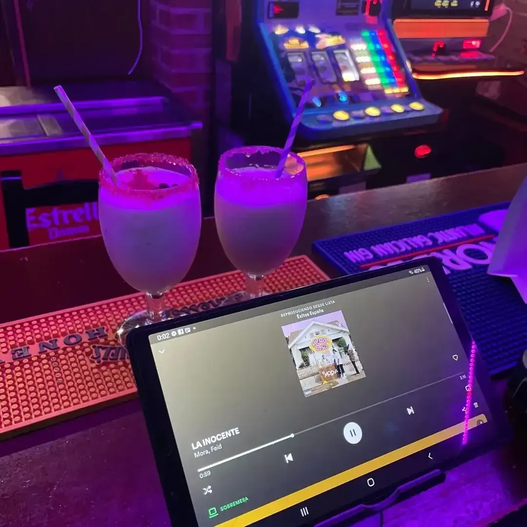 Dos cócteles y una tablet reproduciendo música en una barra, con una máquina tragamonedas de fondo.