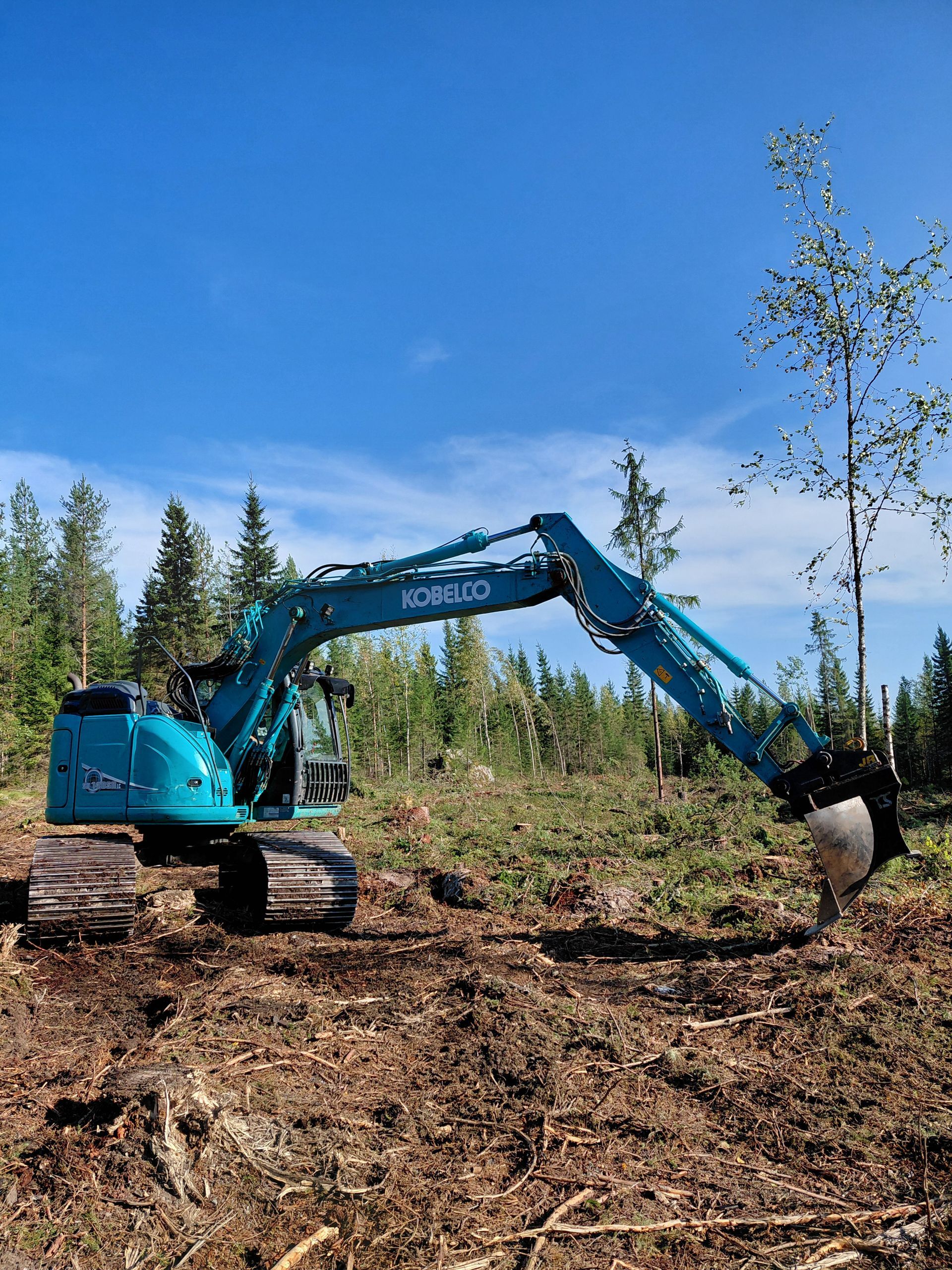 Sininen kaivinkone raivatulla metsäalueella kirkkaan sinisen taivaan alla.
