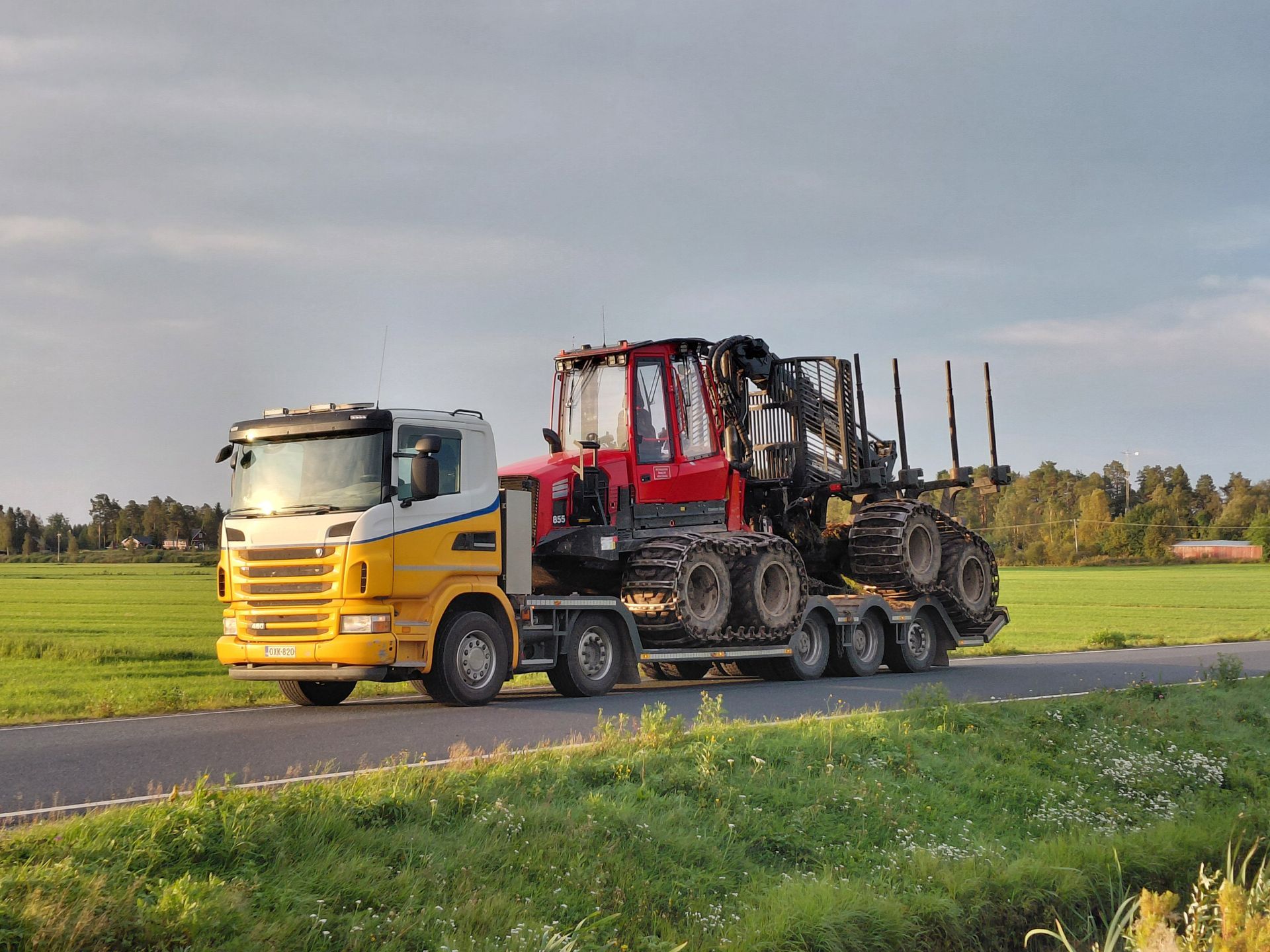 Keltavalkoinen kuorma-auto vetää punaista metsäkonetta perävaunulla tietä pitkin viheralueella.