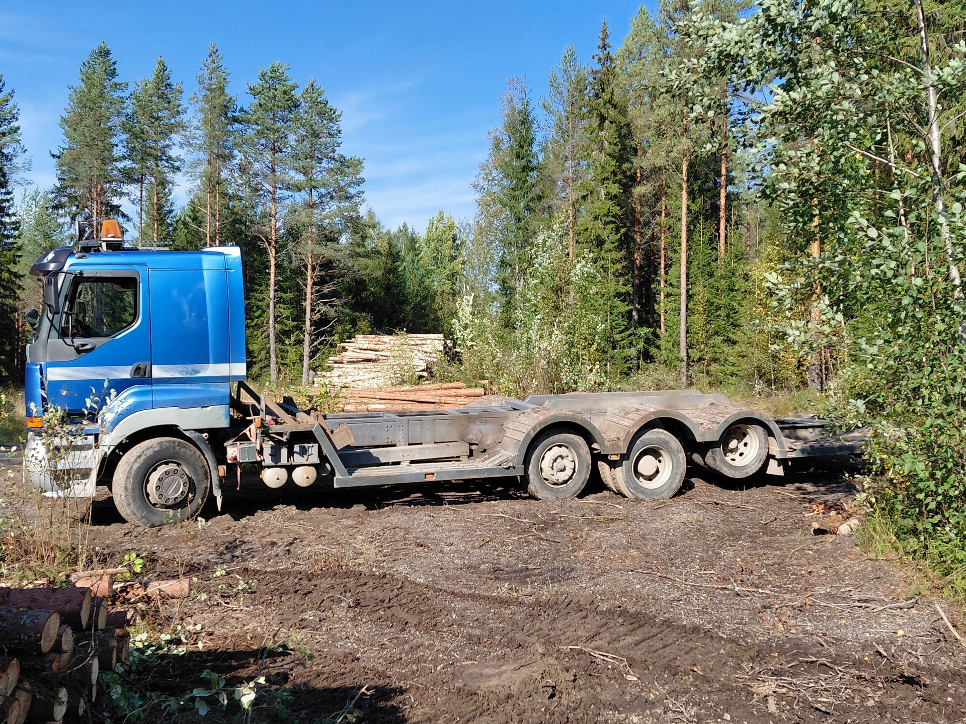 Sininen puoliperävaunu ja lavetti mutaisella metsätiellä. Puita taustalla aurinkoisena päivänä.