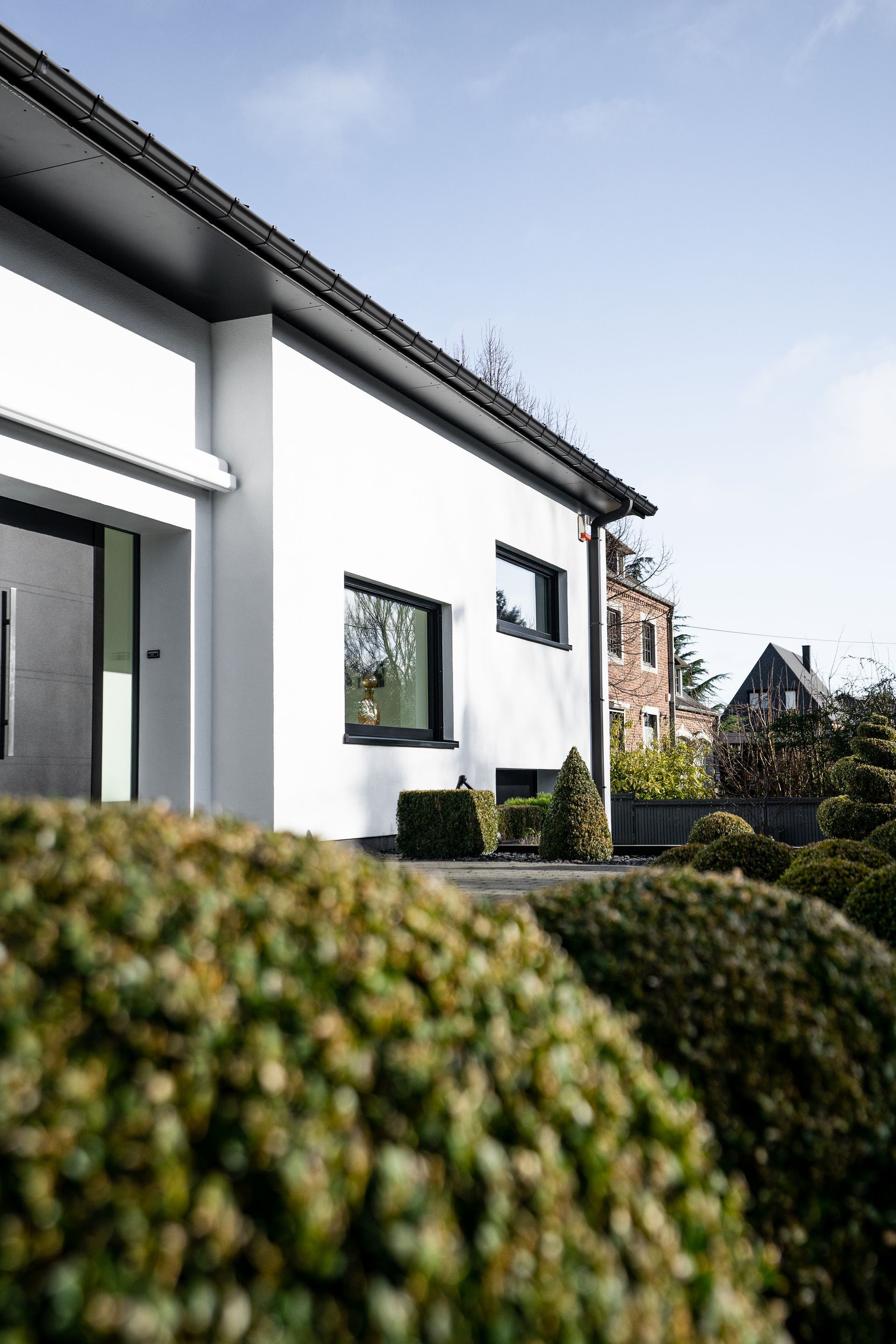 Maison blanche moderne avec garnitures noires, petites fenêtres et arbustes verts devant.