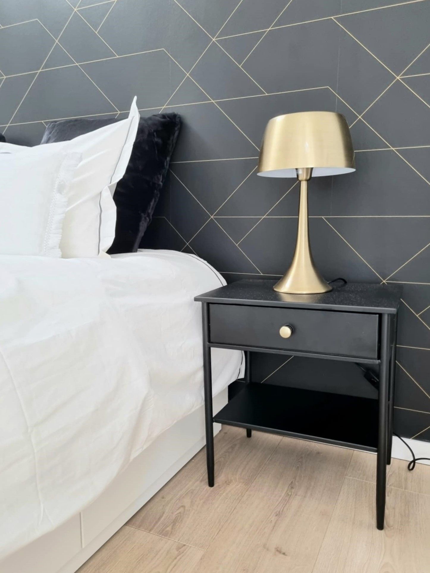 Table de chevet noire avec lampe dorée à côté du lit avec literie blanche et oreiller noir contre un mur à motifs géométriques.