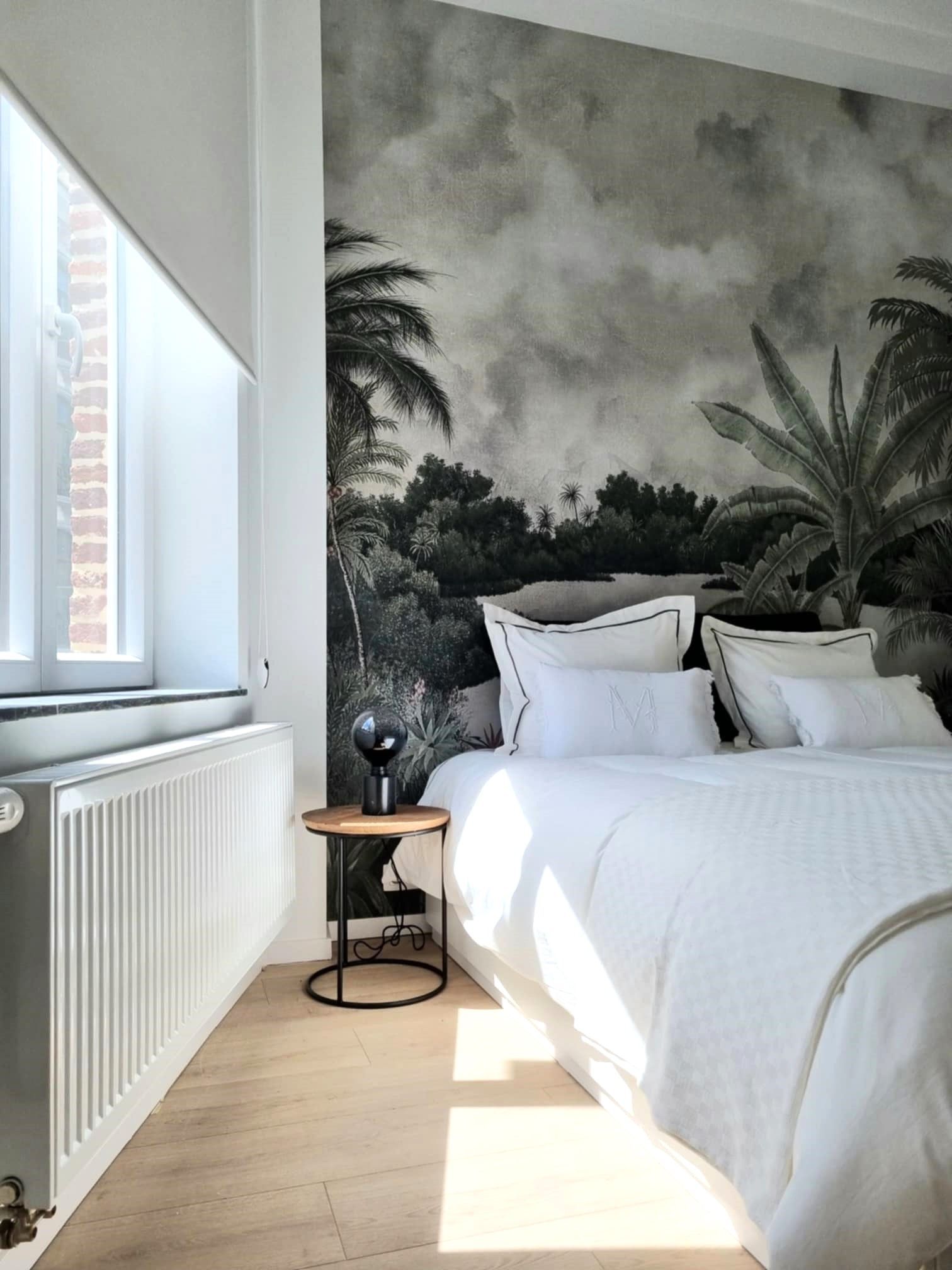 Chambre avec un lit blanc, une peinture murale tropicale, une petite table et une fenêtre avec lumière du soleil.