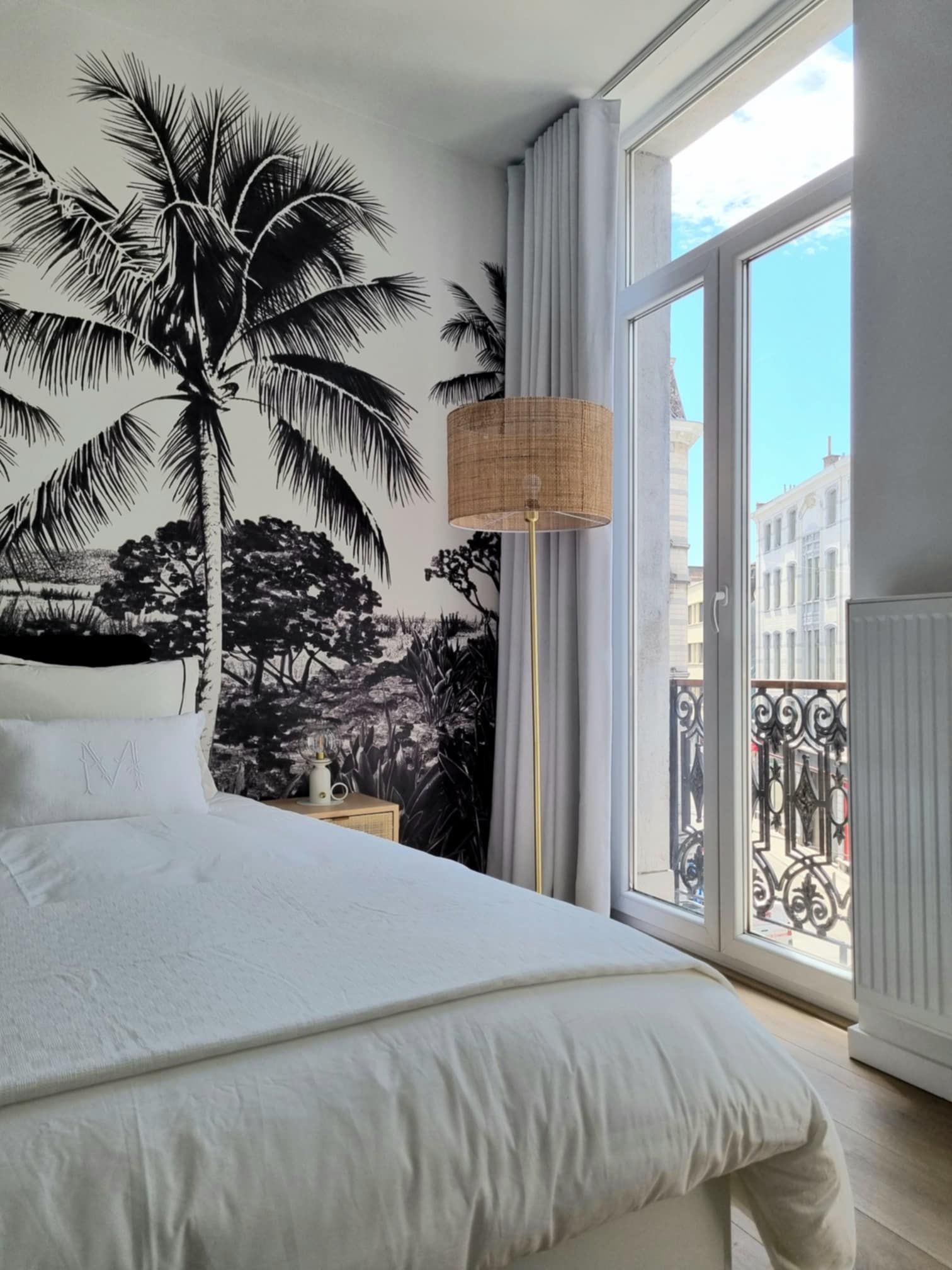 Chambre avec fresque murale de palmier noir et blanc, lit blanc et portes-fenêtres ensoleillées.