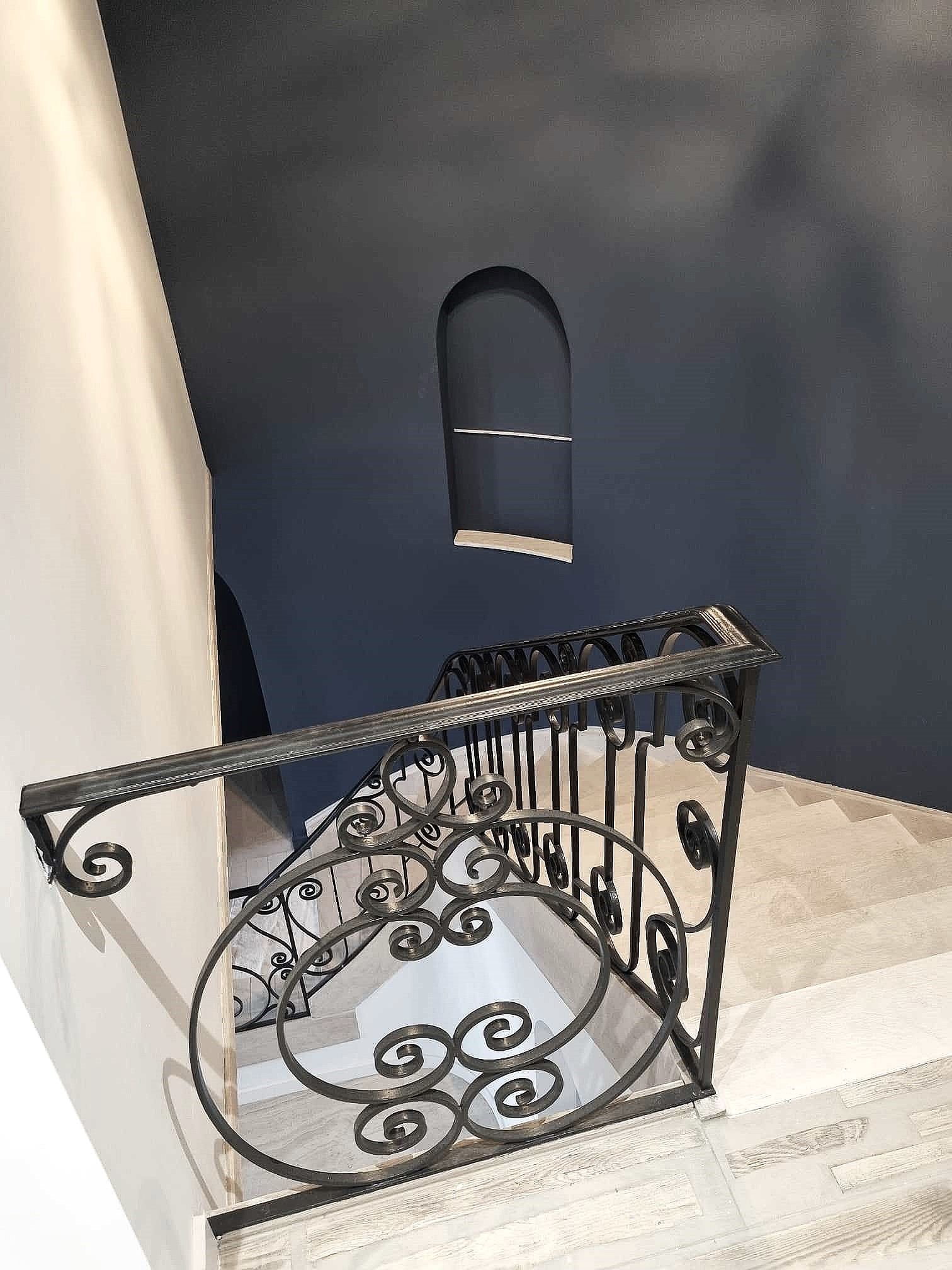 Rampe en fer forgé noir sur un escalier avec un mur bleu et un petit insert en arc.