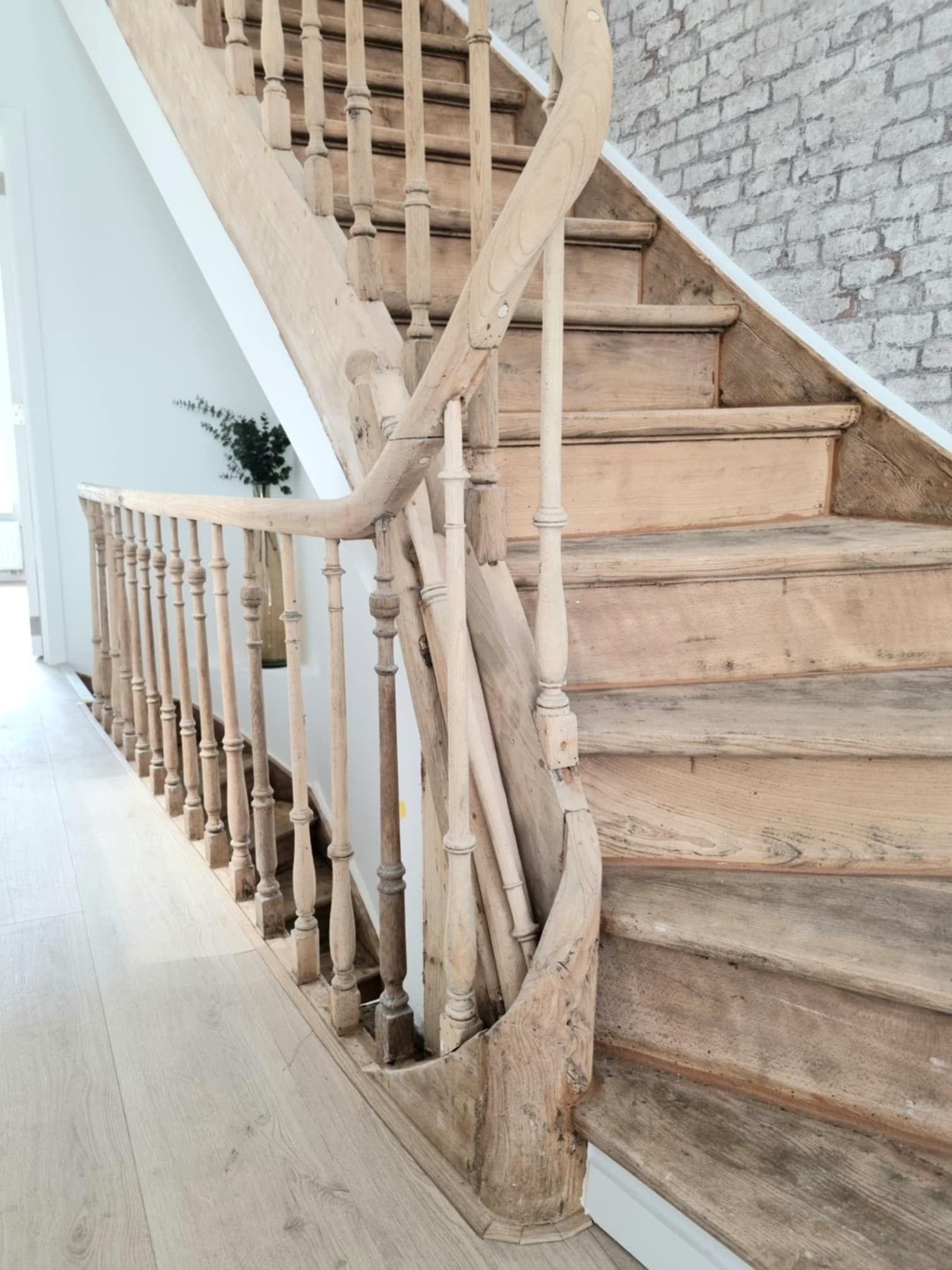 Escalier en bois avec marches usées et rampe ornée, menant vers le haut près d'un mur de briques.