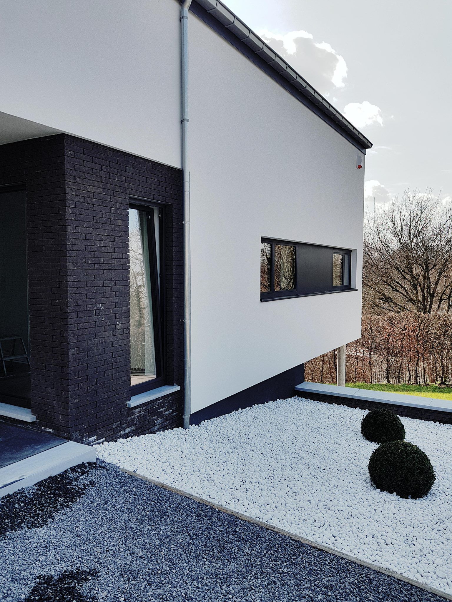 Maison blanche moderne avec des accents de briques foncées, de petites fenêtres et un aménagement paysager en pierre blanche.