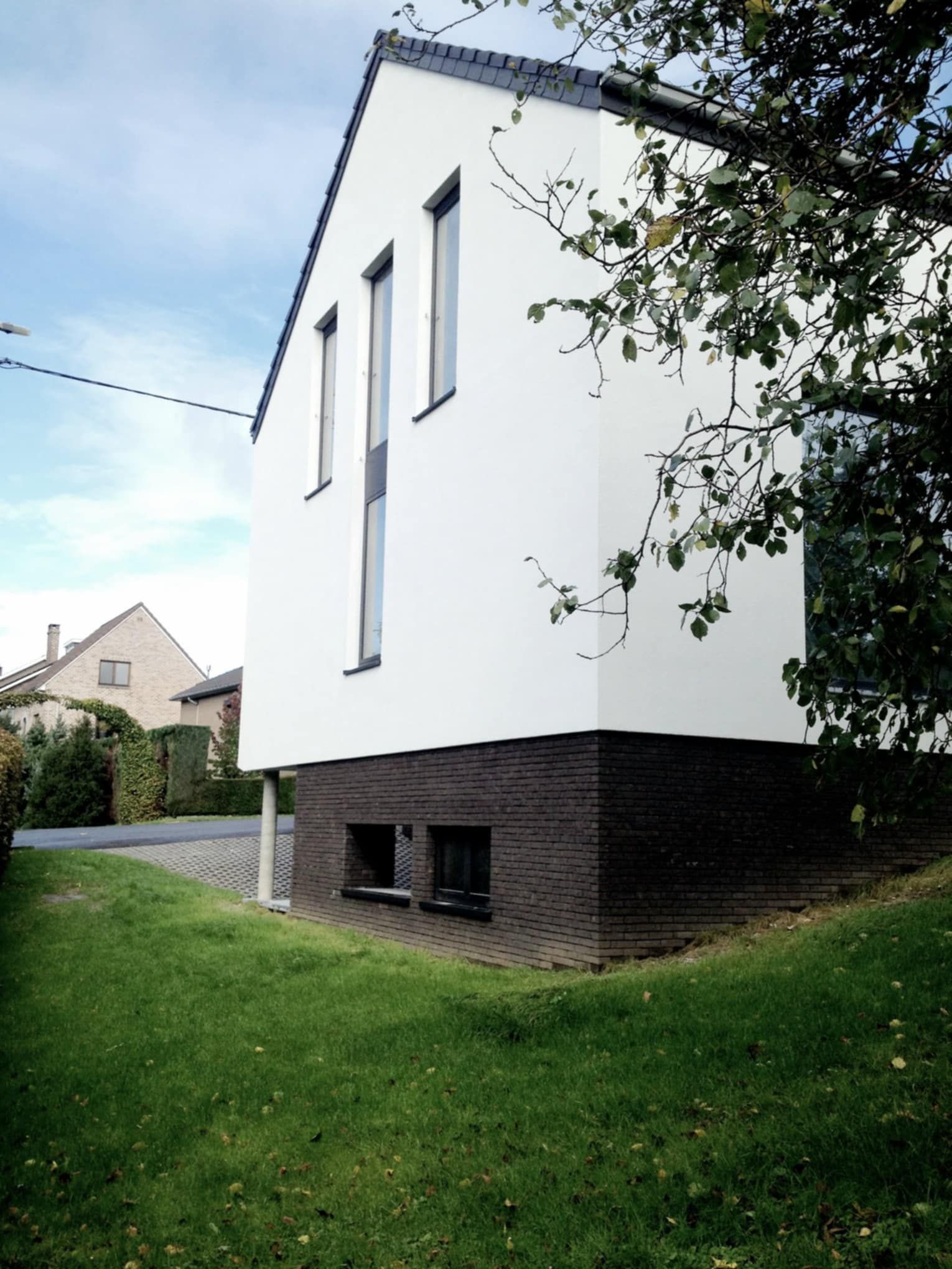 Maison blanche moderne avec base en briques foncées, fenêtres hautes et étroites, pelouse verte.