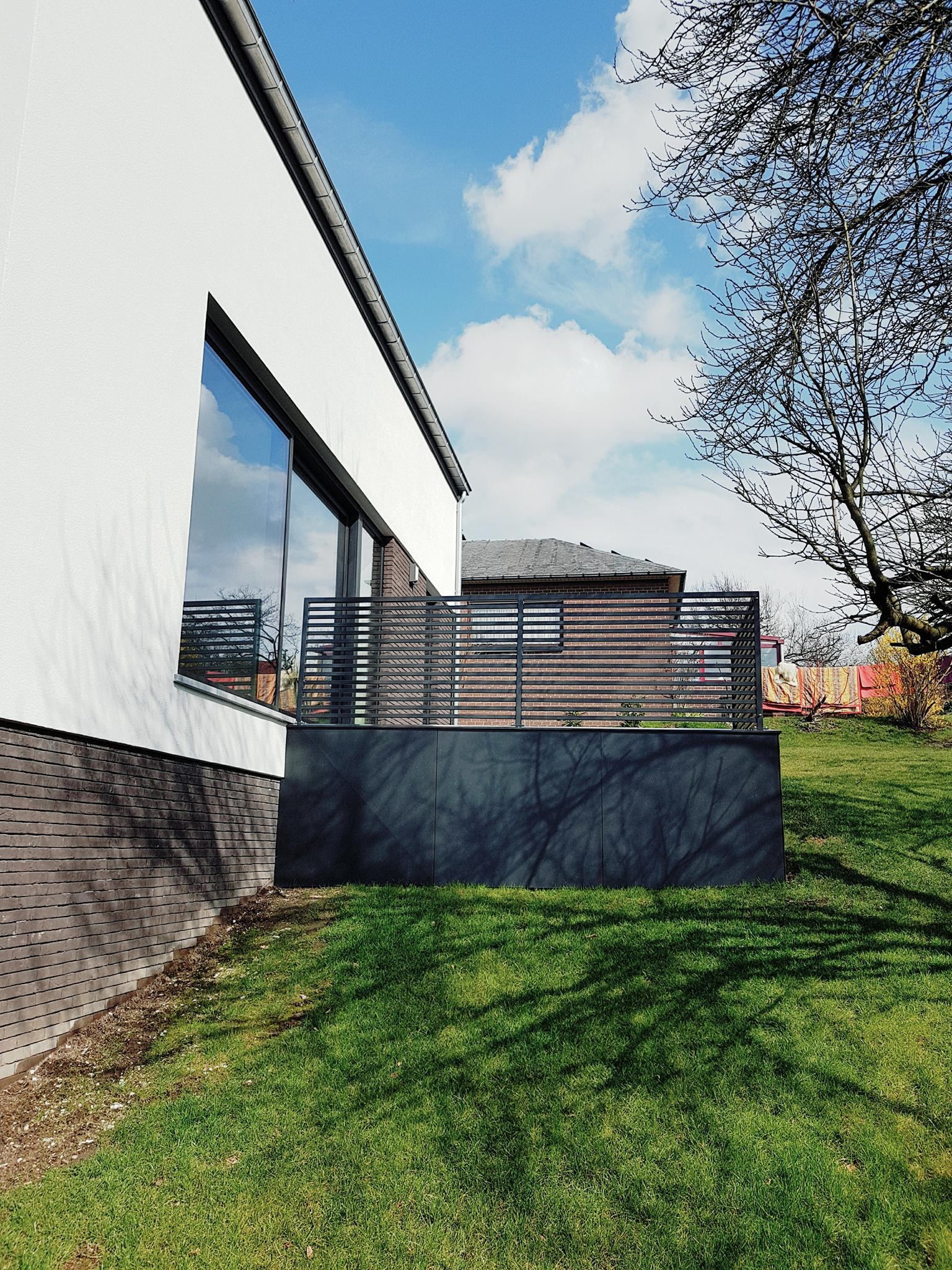 Maison blanche moderne avec une grande fenêtre, une clôture sombre et de l'herbe verte contre un ciel bleu.