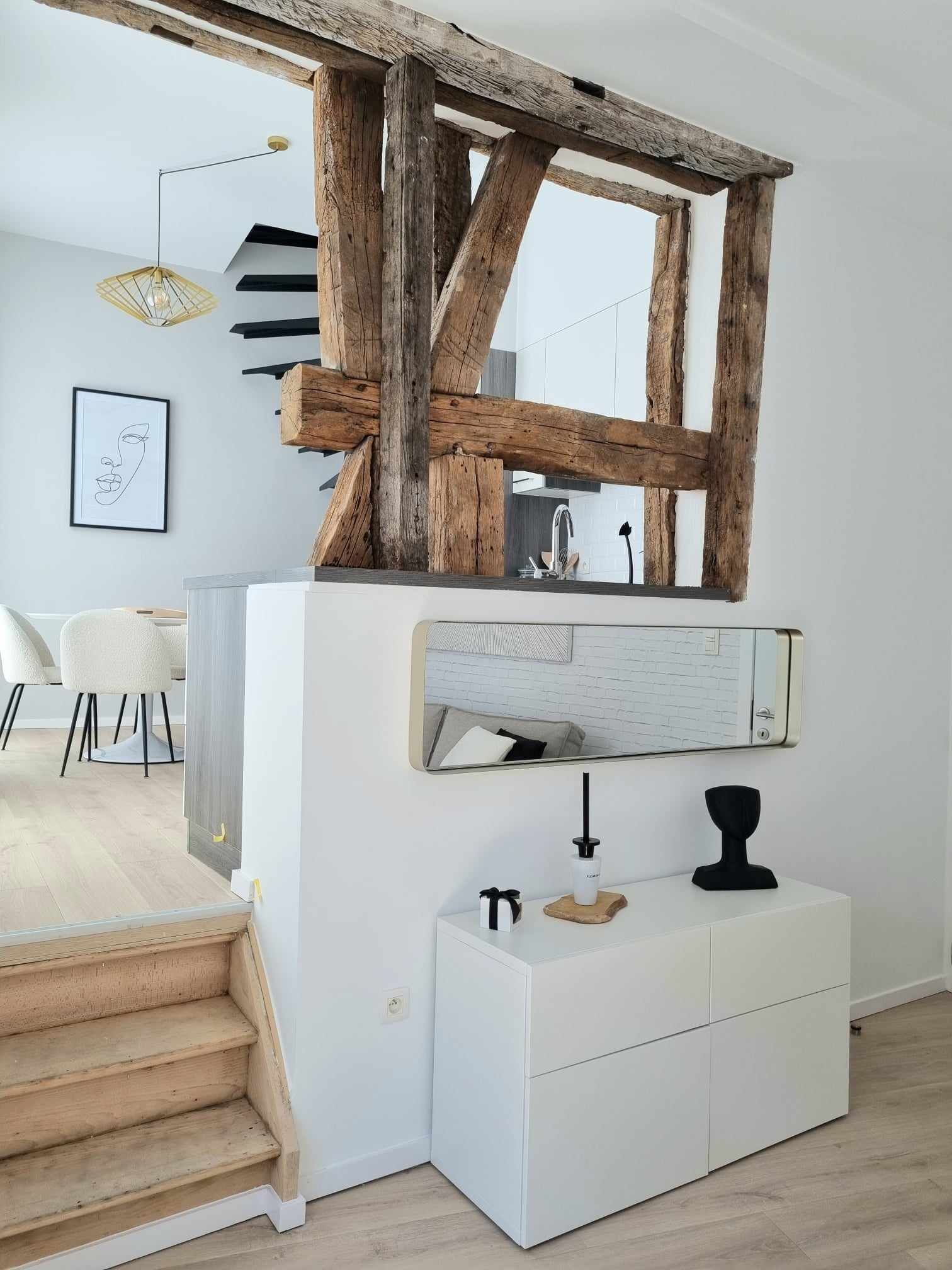 Mur blanc avec poutre en bois rustique, armoire blanche, miroir et un aperçu de la cuisine et des escaliers.
