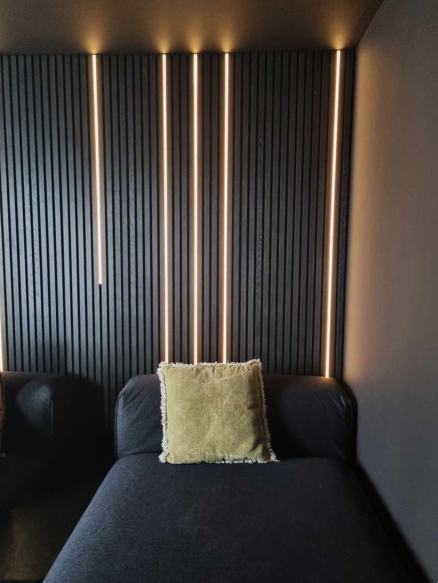 Mur cannelé gris foncé avec éclairage vertical doré au-dessus d'une chaise longue noire avec un coussin doré.