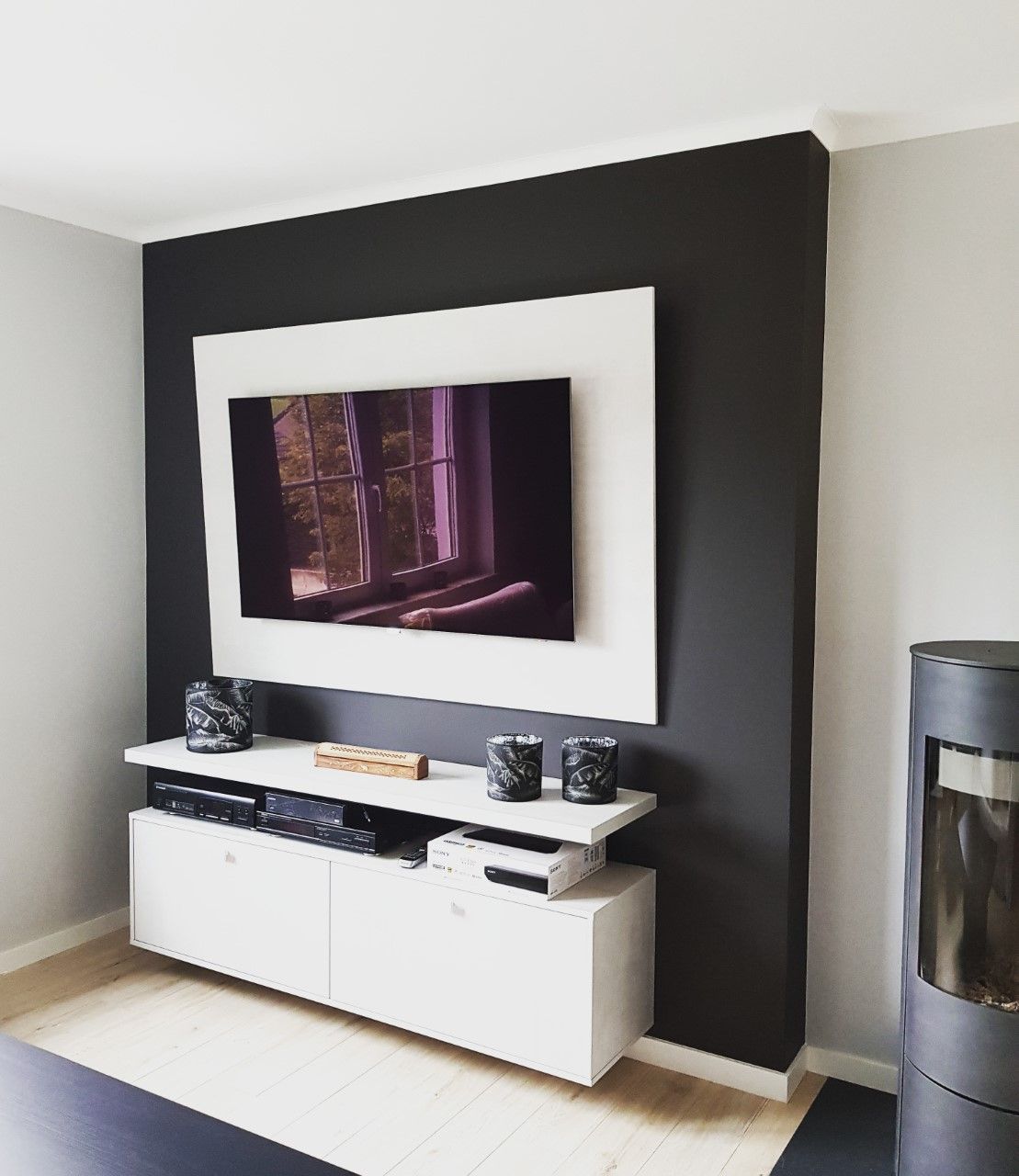 Mur noir avec cadre blanc autour d'un téléviseur, au-dessus d'une console multimédia blanche, à côté d'un poêle gris.