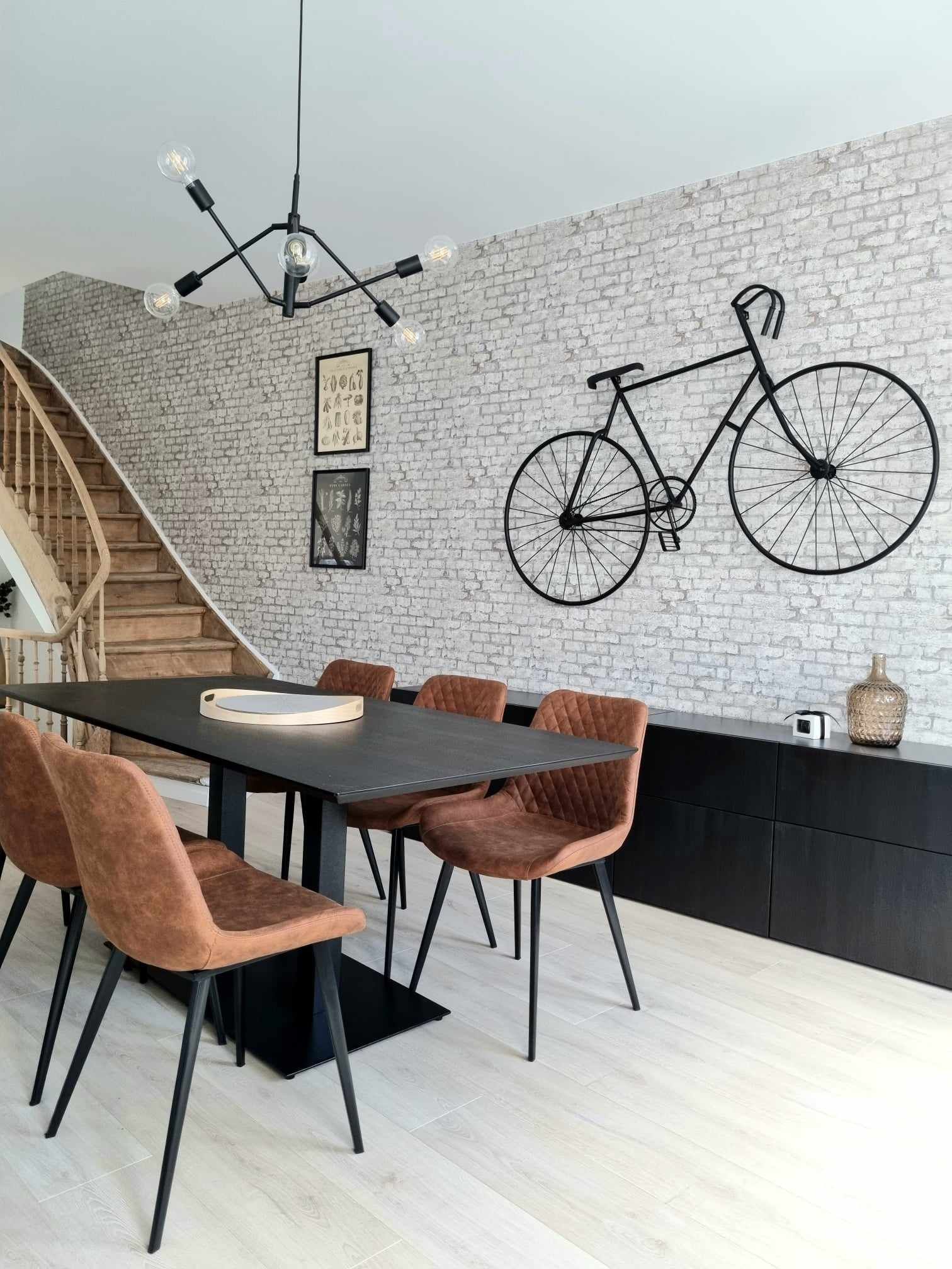Salle à manger : table noire, chaises marron, décoration murale vélo, briques apparentes, escalier en bois.