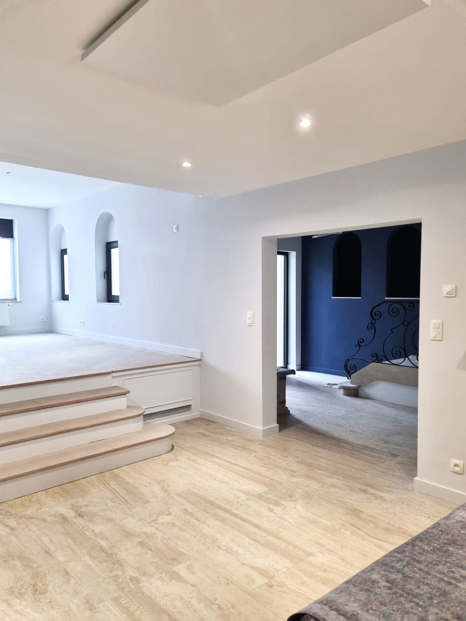 Un intérieur avec parquet clair et escalier menant à une plateforme surélevée perchée de fenêtres en arc. Un mur bleu est visible.