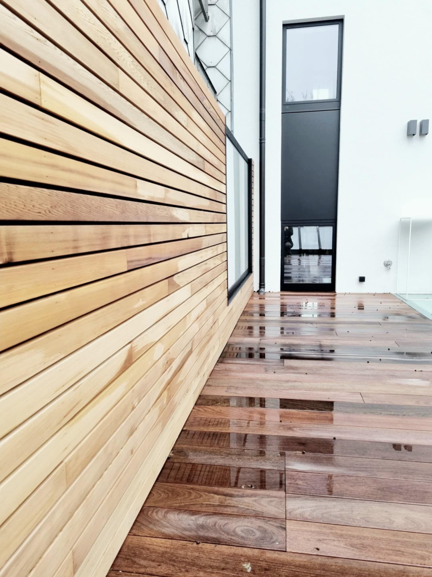 Terrasse en bois et mur de lattes de bois horizontales à côté d'un bâtiment blanc avec des fenêtres.